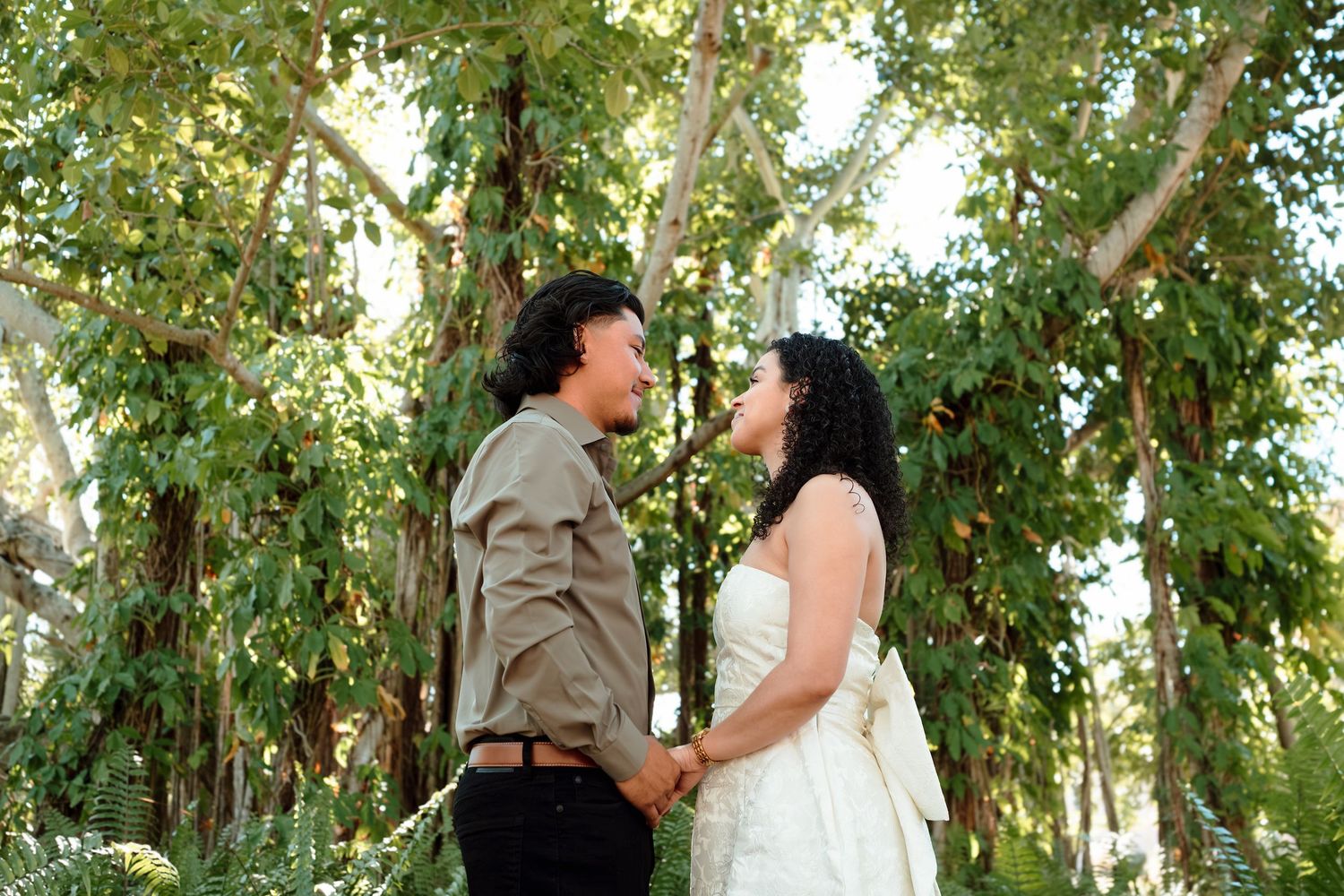 A couple holds hands and gazes at each other lovingly outdoors, surrounded by lush green trees in a romantic wedding setting.