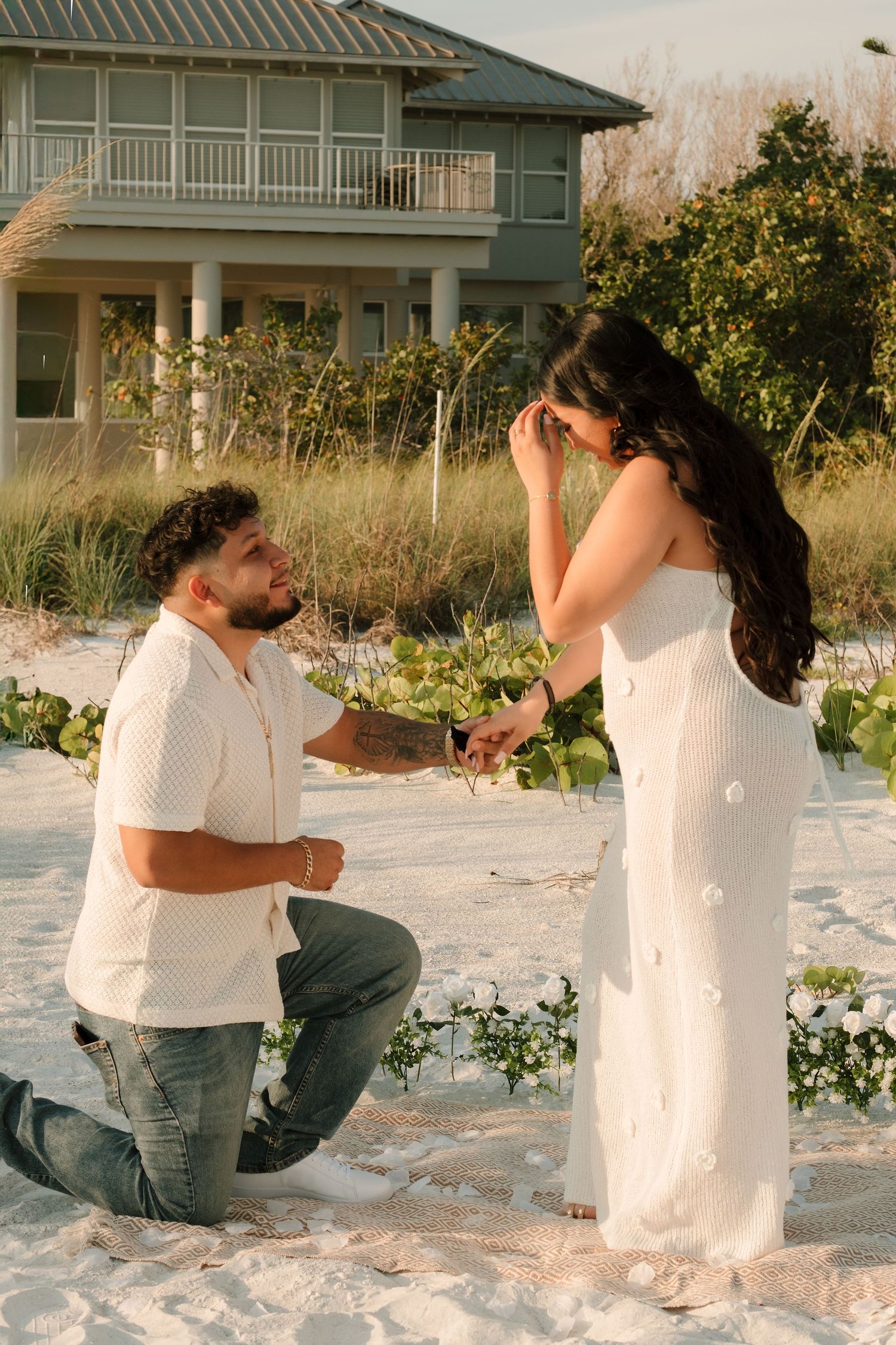 Man proposes on one knee to woman in white dress on sandy beach near coastal home at sunset.