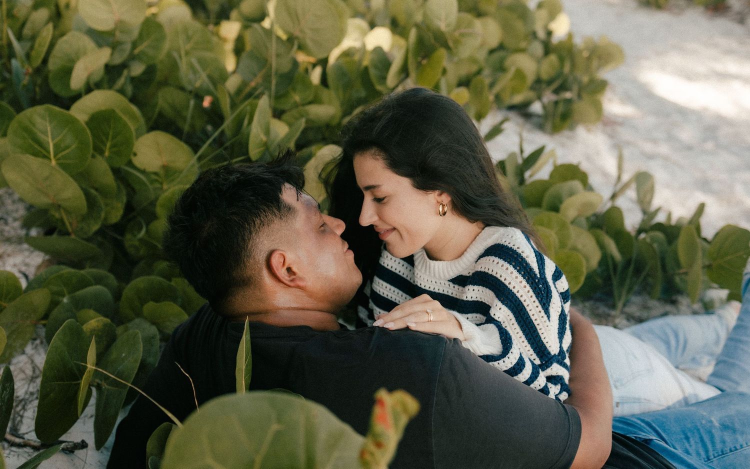 Romantic couple sharing an intimate moment together outdoors near lush green tropical plants on a sandy beach.