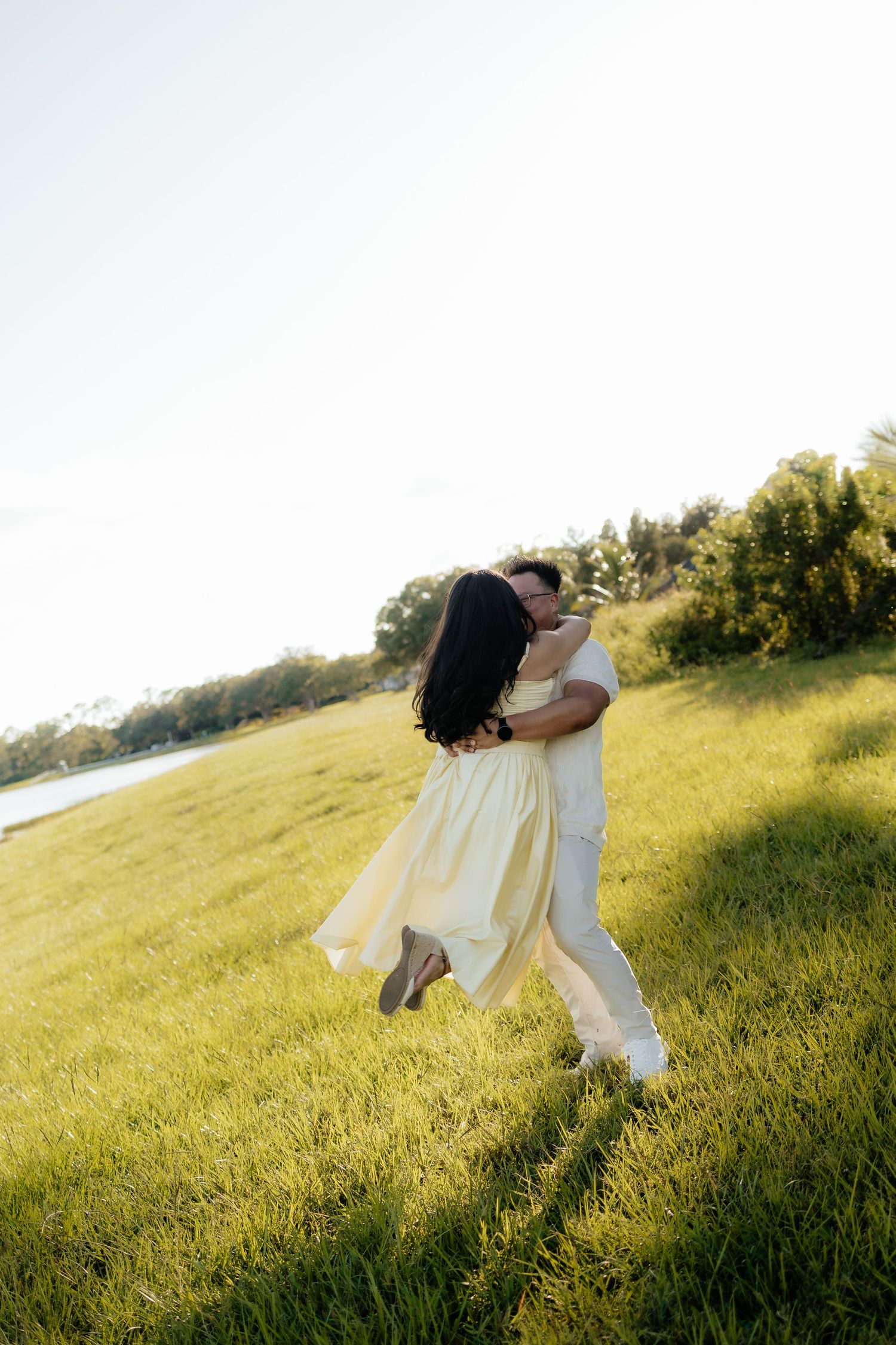 A couple embraces and spins joyfully in a sunlit green field near a lake, the woman in a flowing yellow dress.