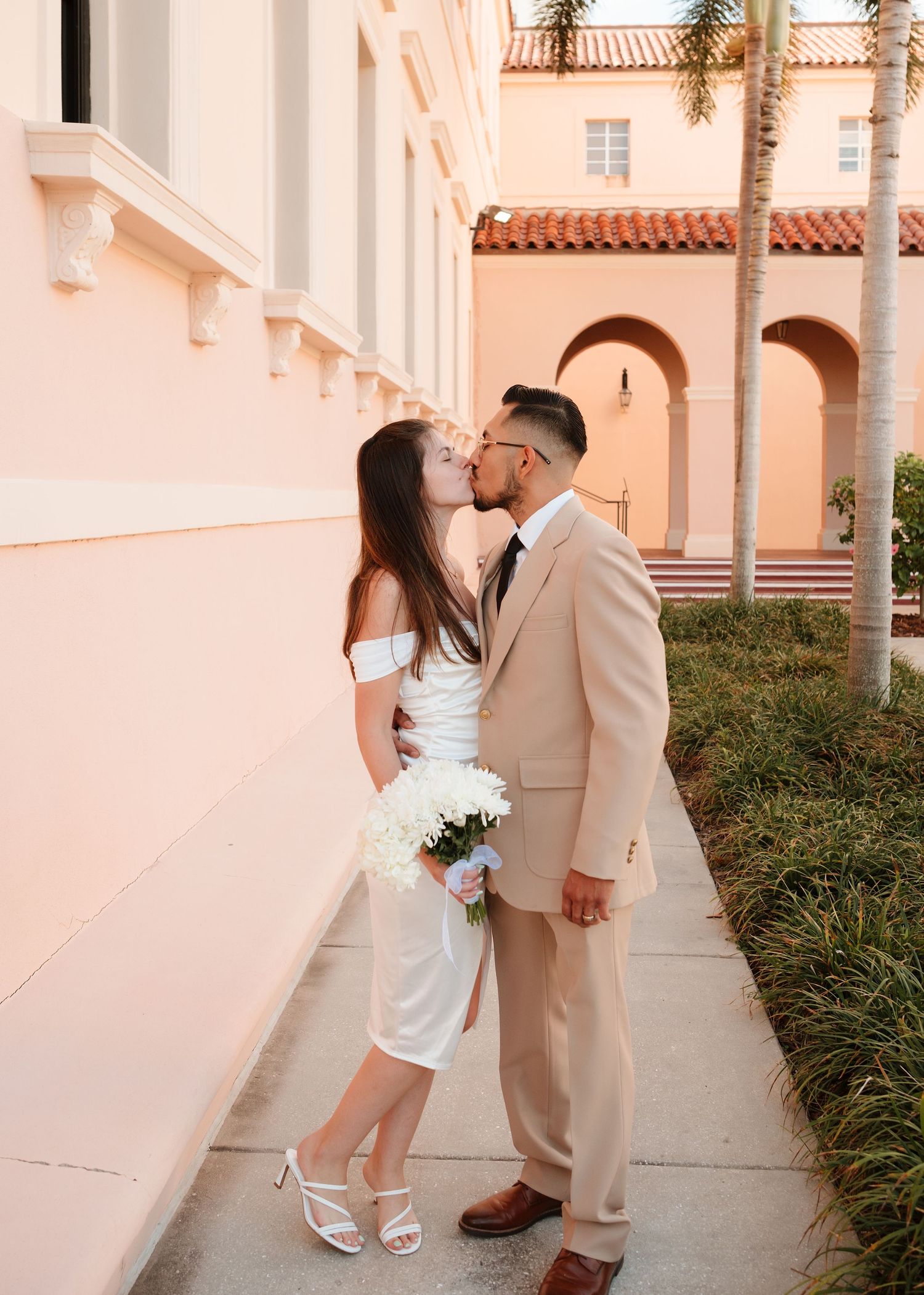 Newlywed couple kissing outside a pink building, bride in white dress holding bouquet, groom in tan suit.
