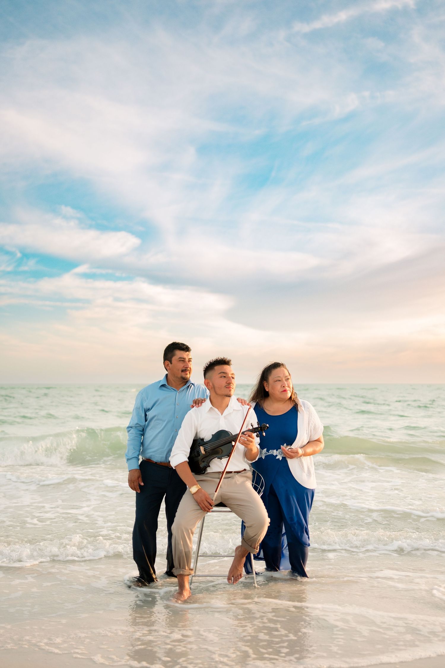 Family of three poses on a beach at sunset, with son playing violin while standing in shallow ocean waves.