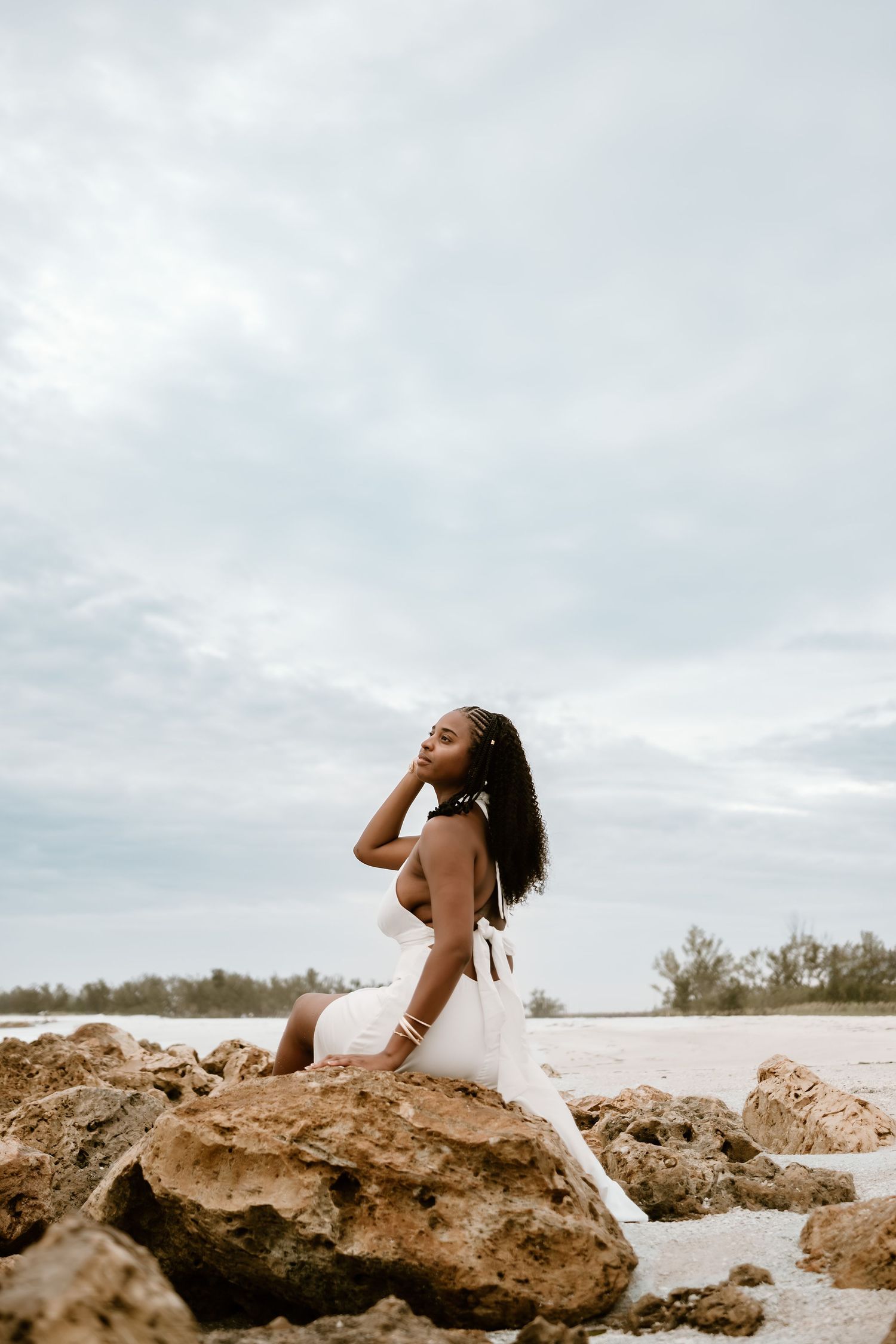 A figure in a white dress poses gracefully on coastal rocks against a cloudy sky at the beach.