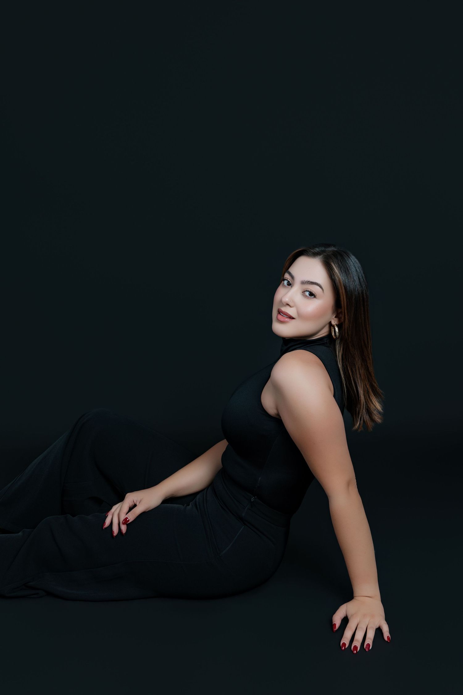 Woman in black outfit poses gracefully on floor against dark background, wearing gold earrings with red nails.