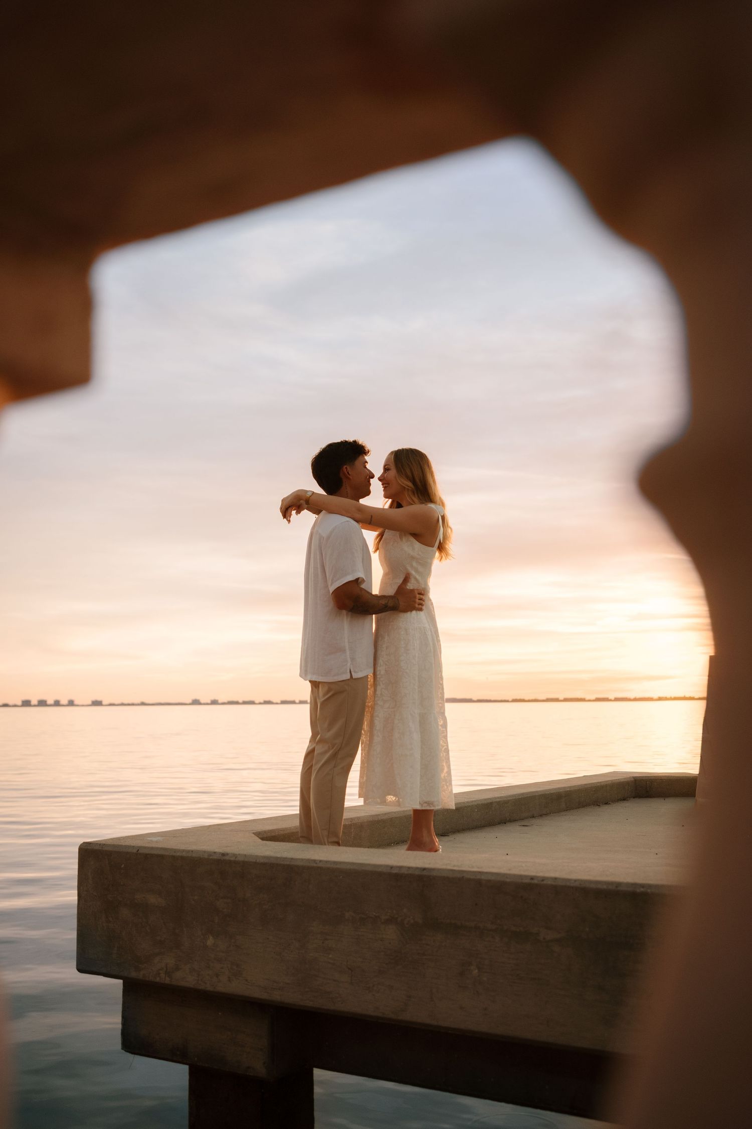 Romantic couple embraces on waterfront pier at golden sunset, framed by stone archway over calm ocean waters.