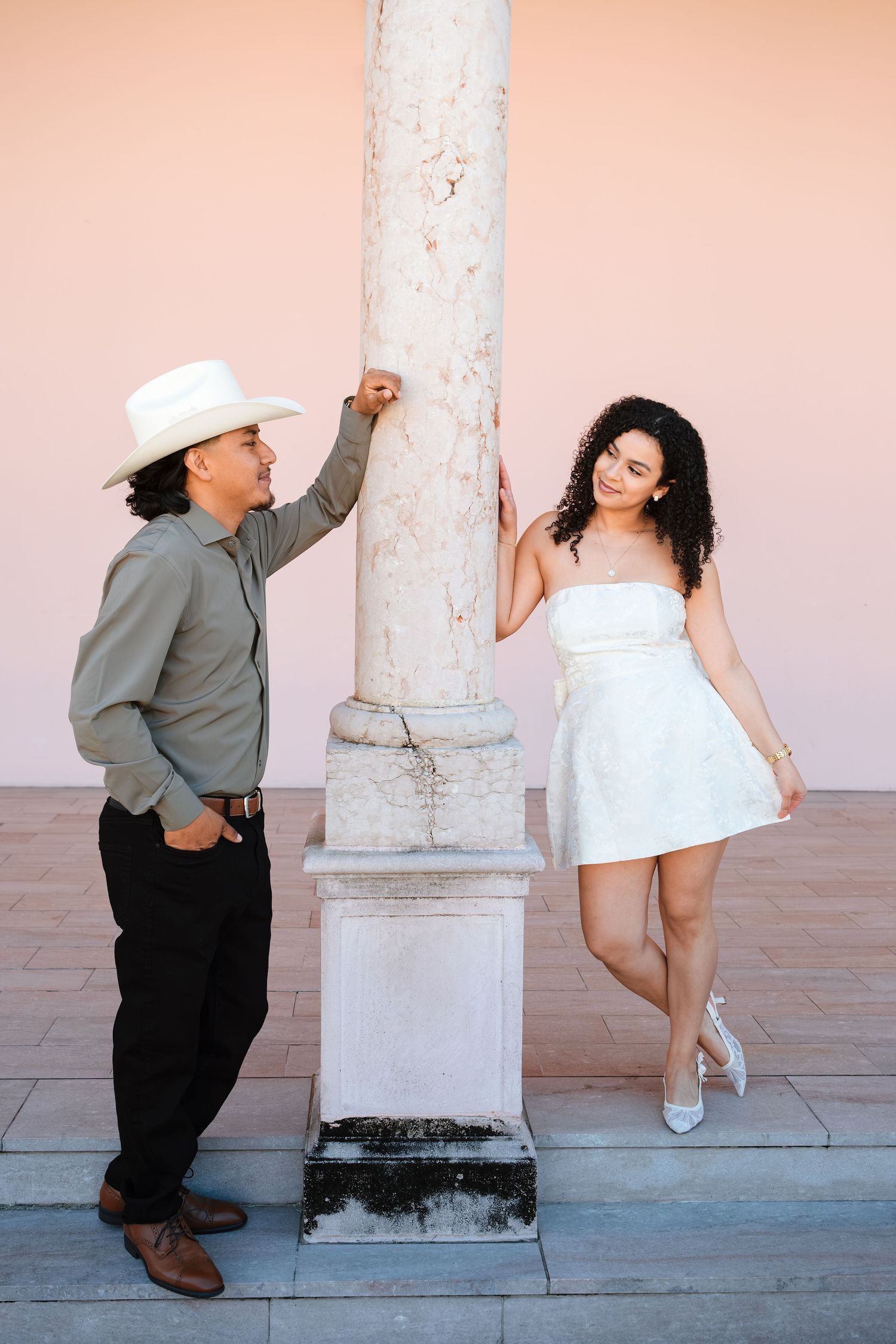 Couple posing by a white column against a pink wall, man in cowboy hat and woman in white dress.