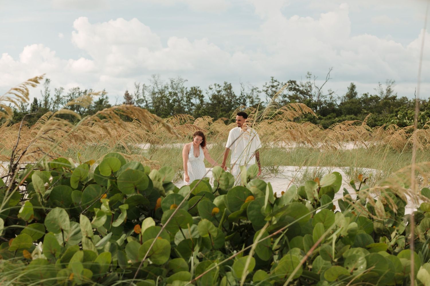 Couple in white outfits walking through lush coastal dunes with tall grass and cacti under a cloudy sky.