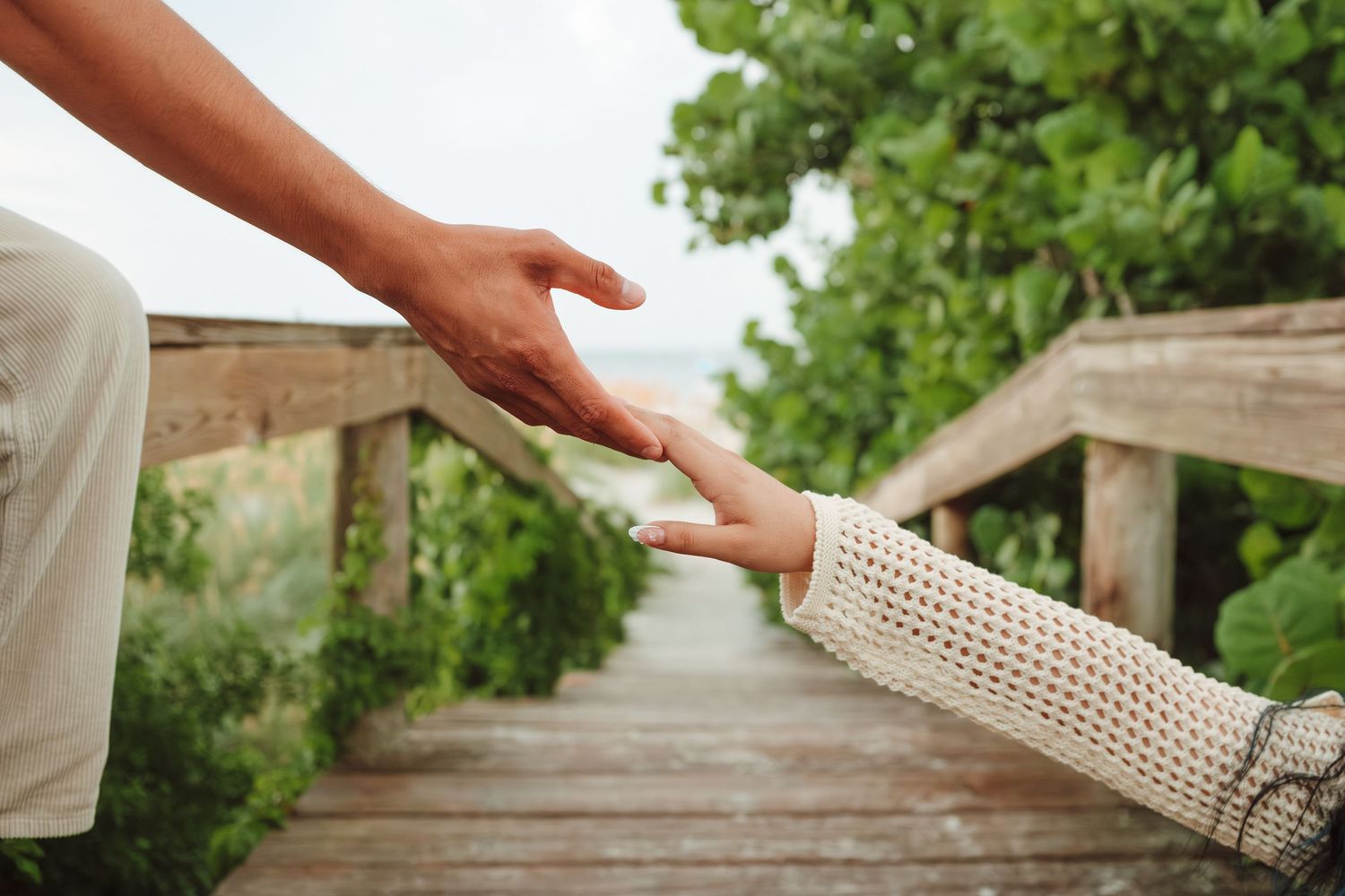 Two people gently holding hands on a wooden boardwalk surrounded by lush green trees near a beach.