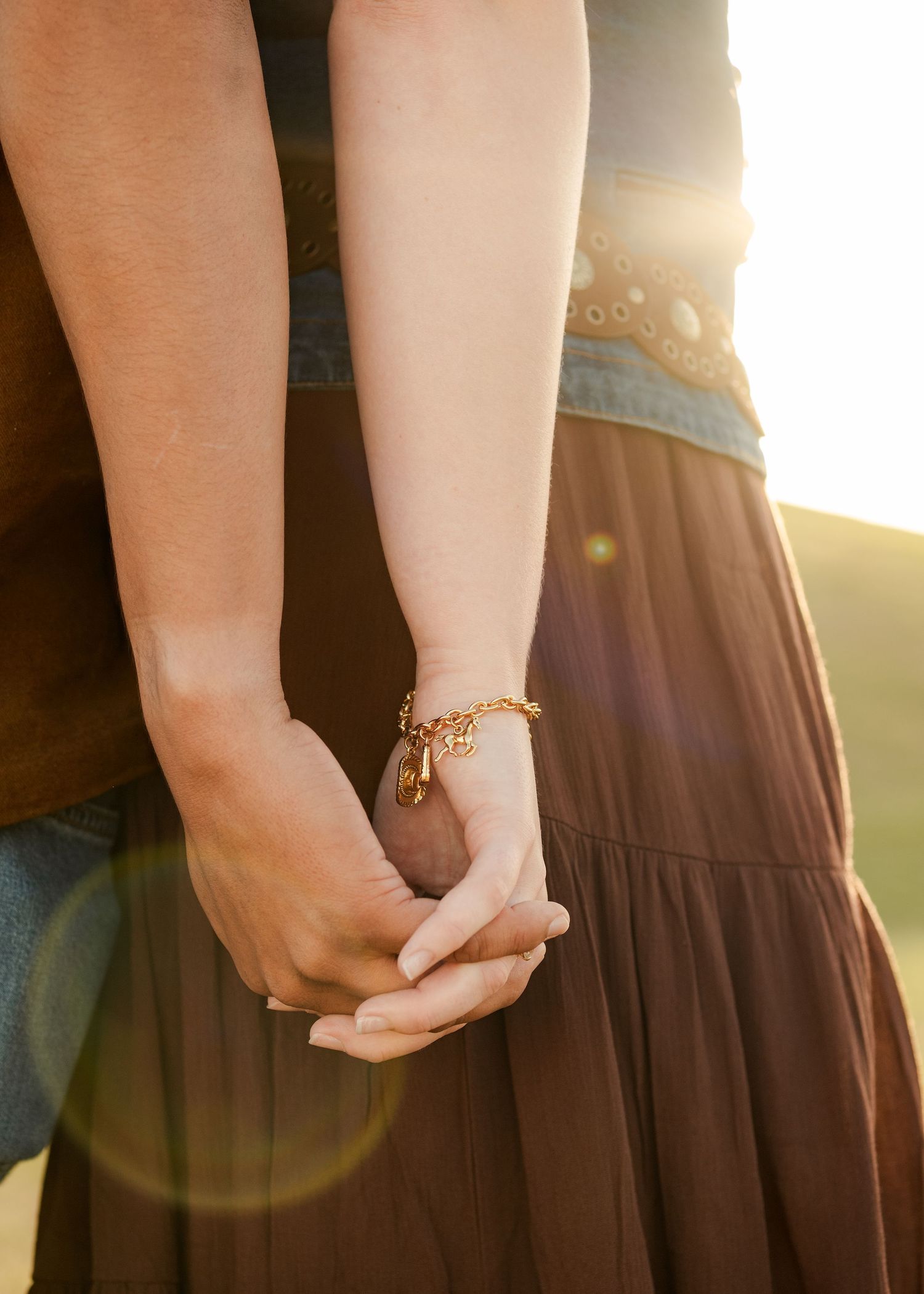 Couple holding hands at golden hour, woman wearing gold charm bracelet, brown maxi skirt and denim top.