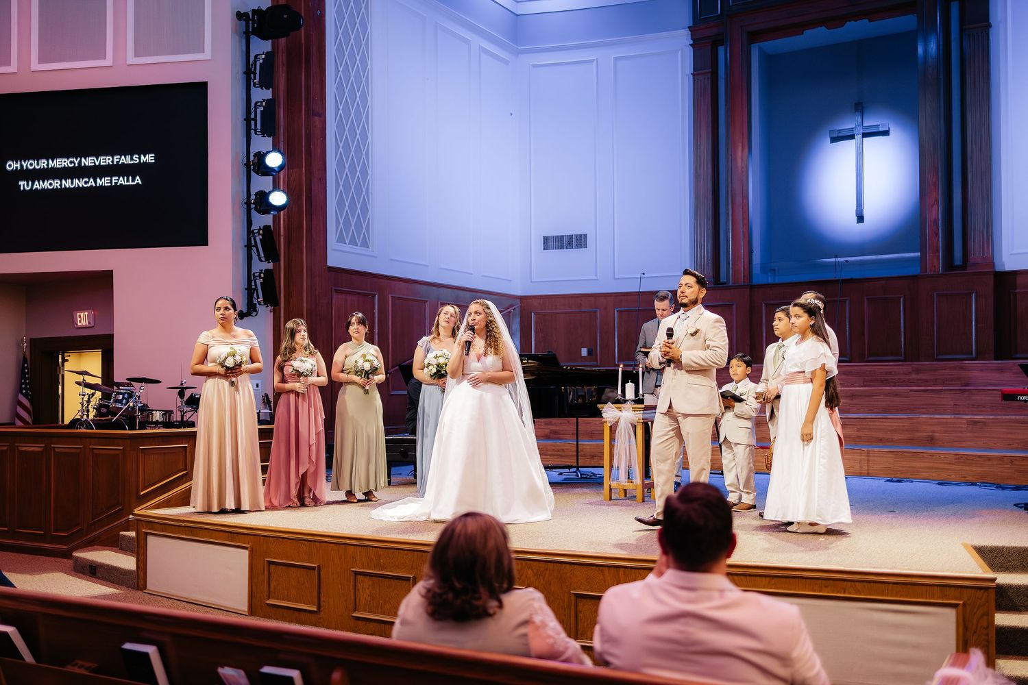 Wedding party stands on church altar stage with cross and bridesmaids in blush gowns during ceremony.