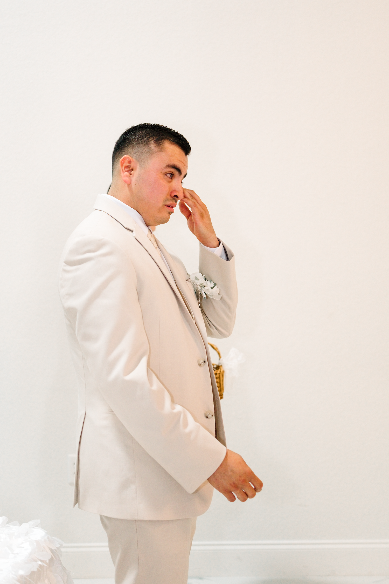 Emotional groom in white suit wiping tears on wedding day, standing against white wall with boutonniere.