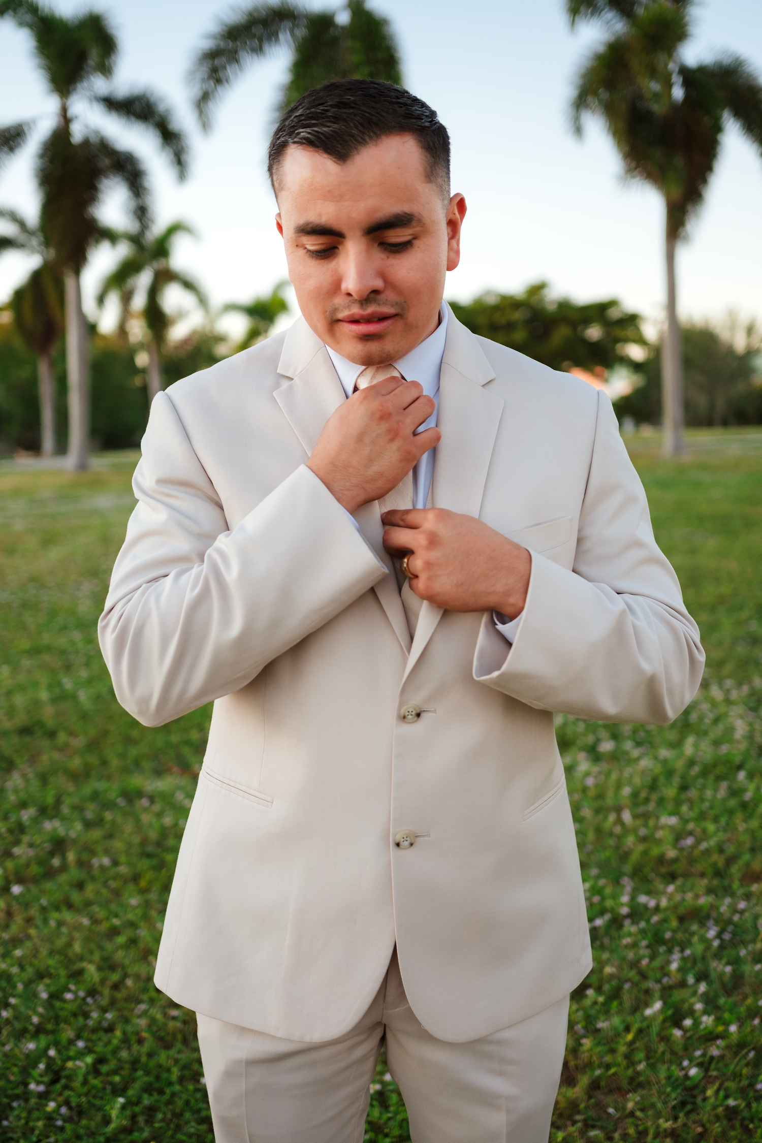 Handsome groom in beige suit adjusts tie outdoors with tropical palm trees in background.