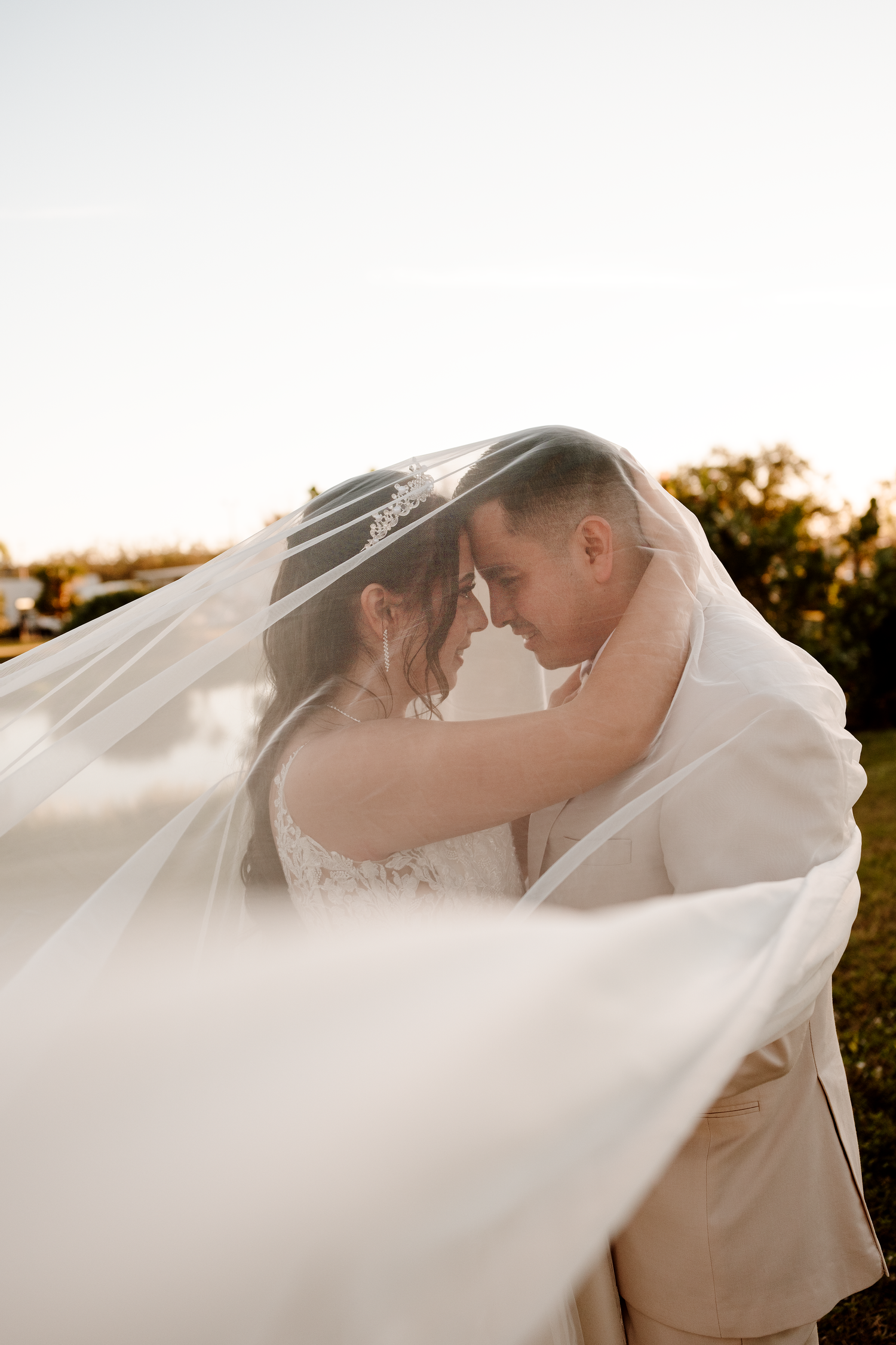 A romantic bride and groom embrace under flowing white veil at sunset outdoors.