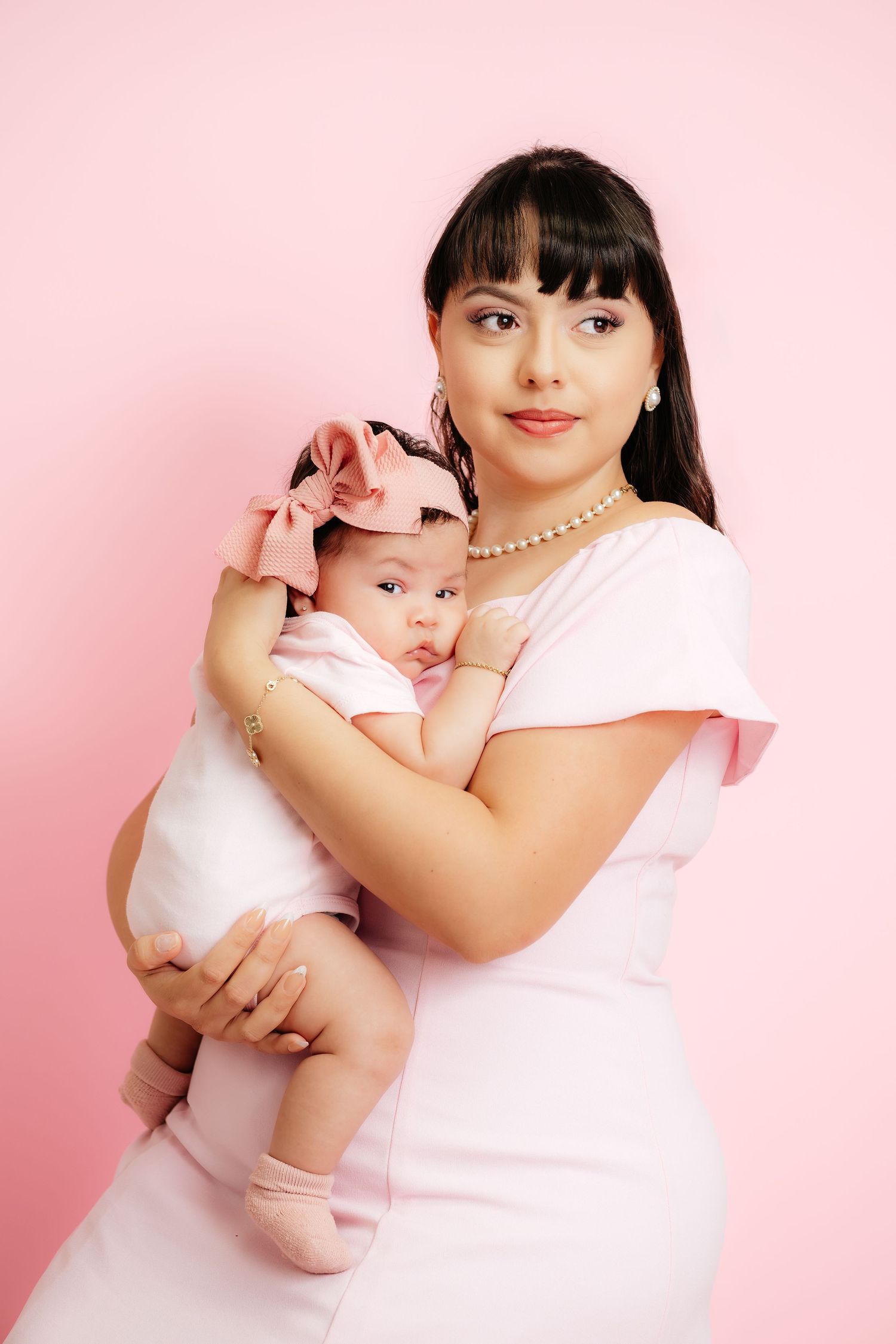 A young mother in a pink dress holds her baby girl with a pink bow headband against a pink background.