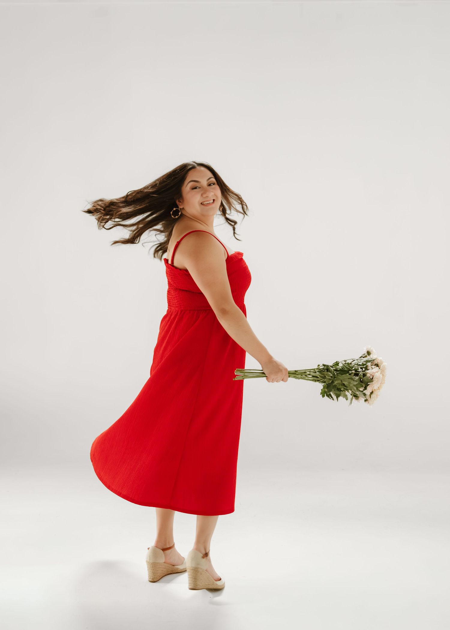 Woman in red midi dress twirls holding white flowers against white background.