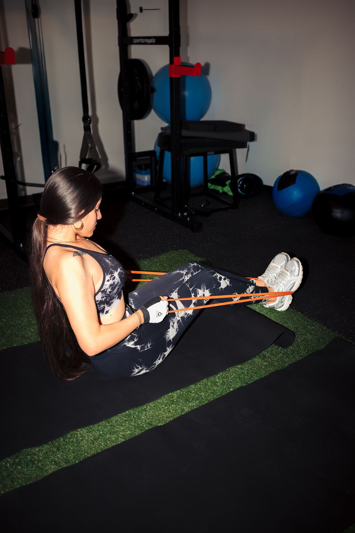 Woman exercising with resistance band on gym floor in black floral activewear, surrounded by fitness equipment.