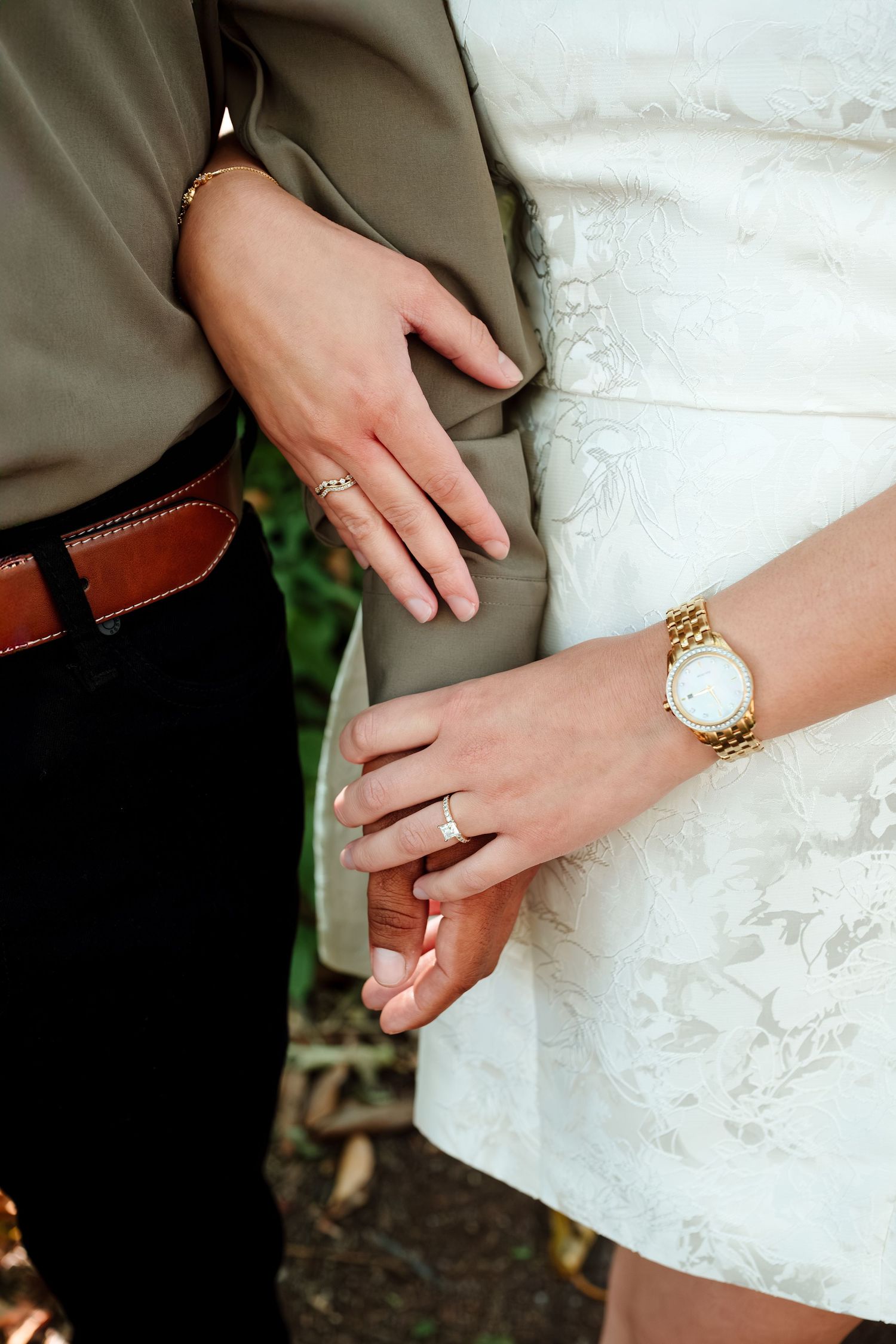 Close-up of a couple holding hands, woman wearing an engagement ring and gold watch, man in black pants with brown belt.