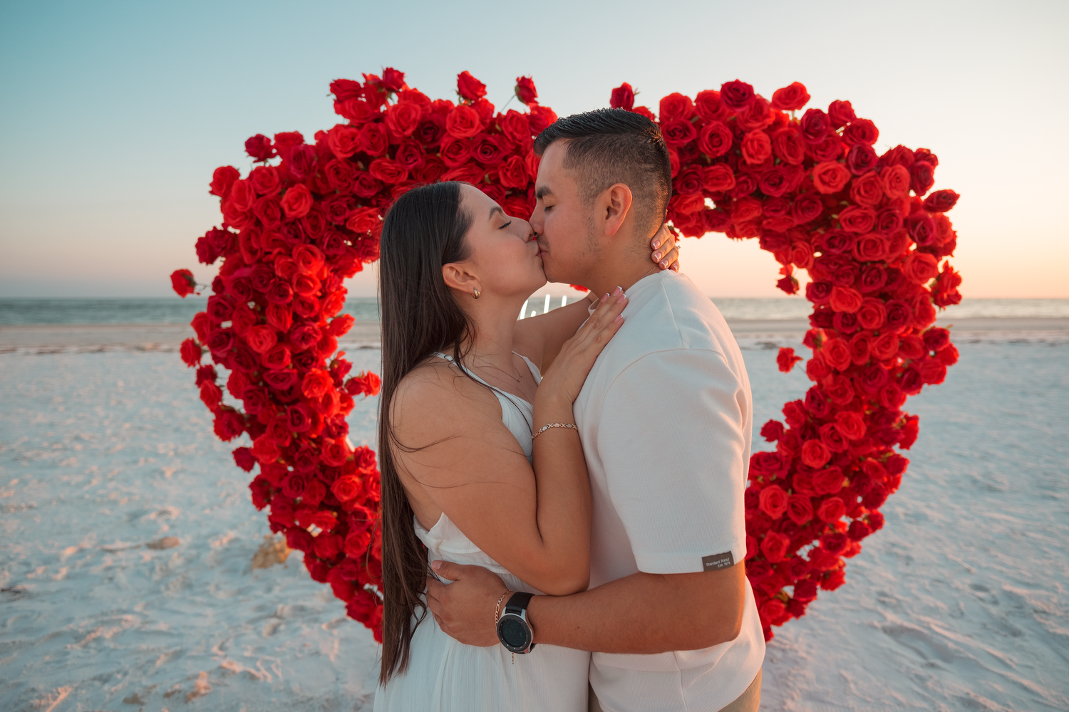Couple kissing on beach in front of red rose heart wreath at sunset.