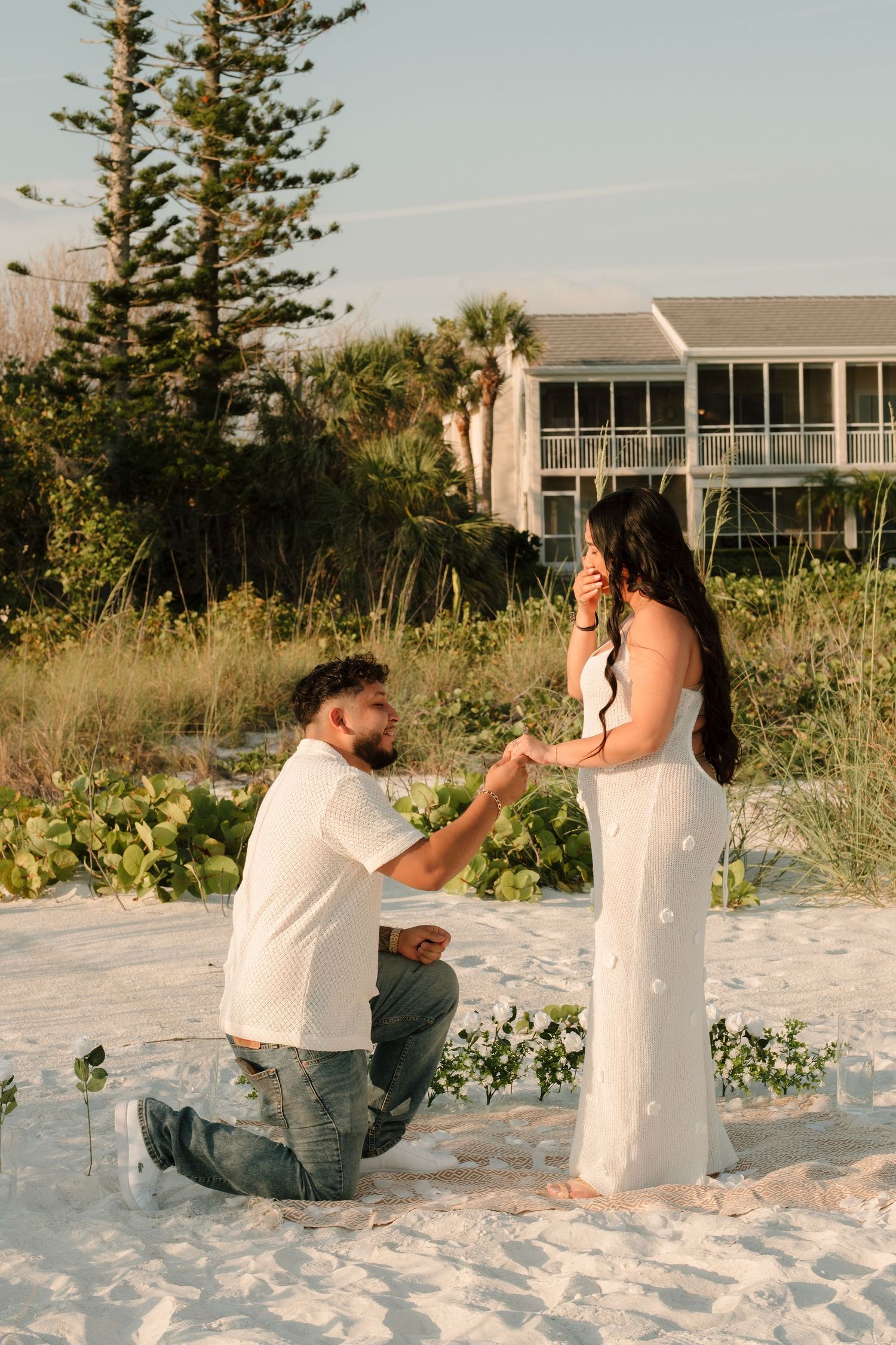 Man kneeling on white sand beach proposing to woman in white dress at golden hour near lush greenery.