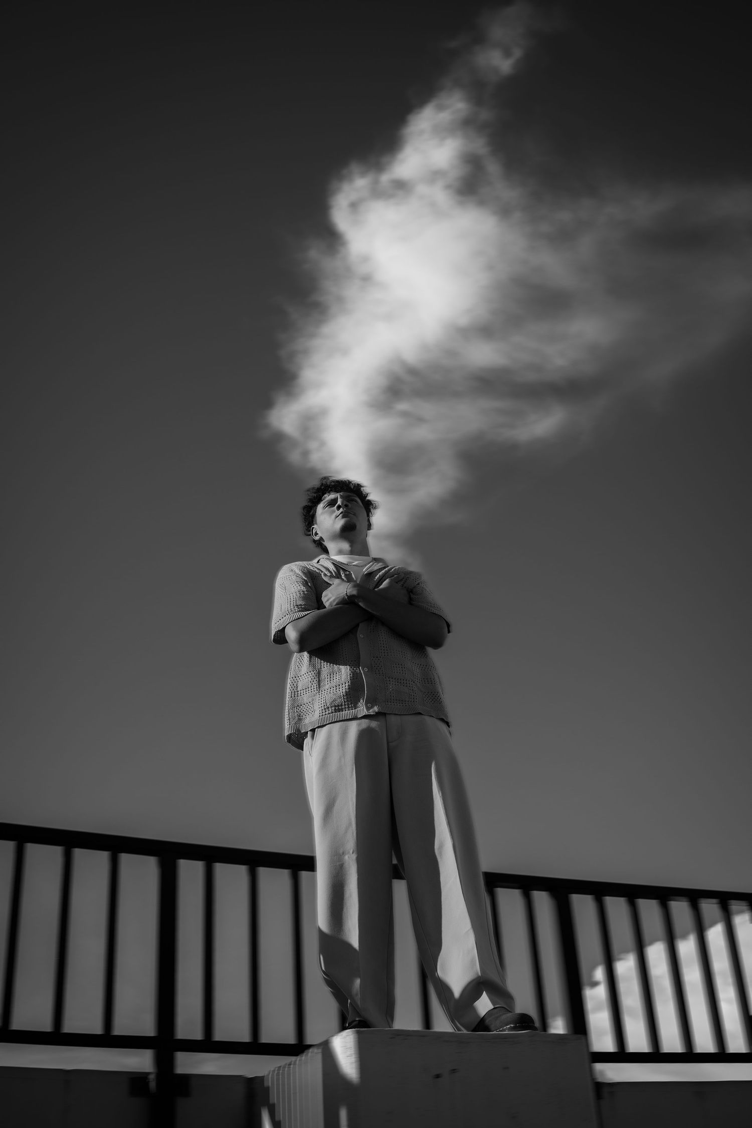 Black and white photo of a person standing on a ledge, arms crossed, exhaling smoke against a dramatic sky.