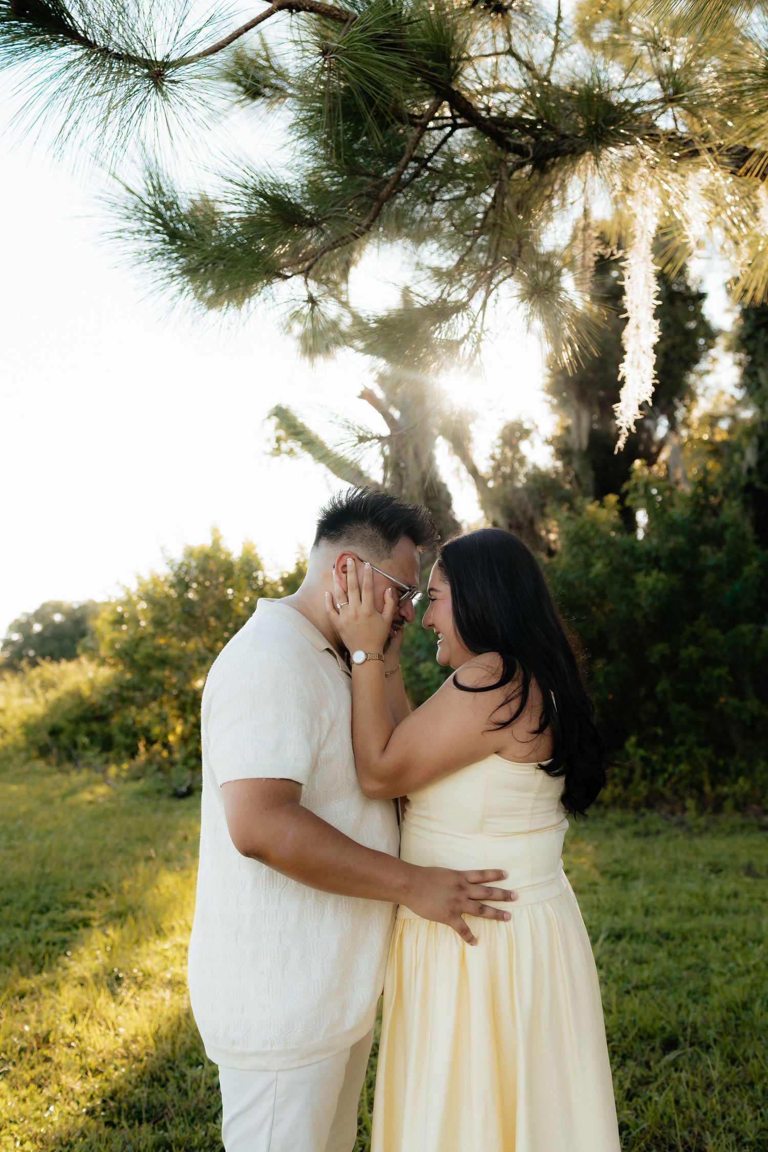 Couple in white and yellow outfits share a romantic moment under a sunlit pine tree in a lush green outdoor setting.