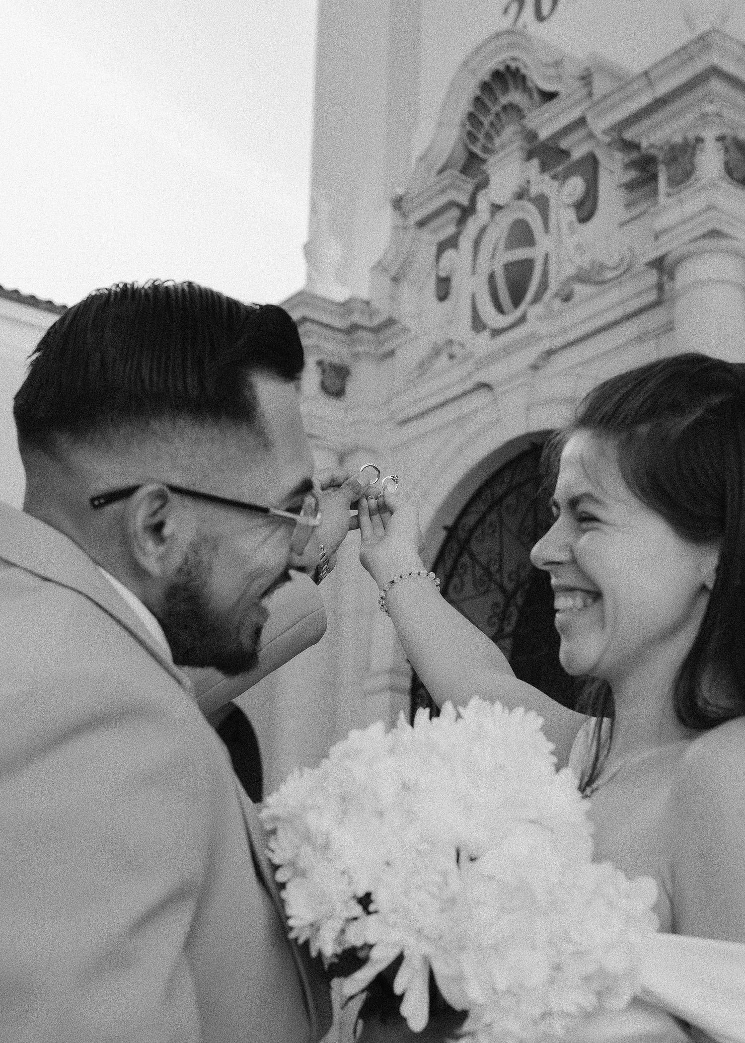 Black and white photo of a smiling bride holding flowers as groom places ring on her finger outside a church.