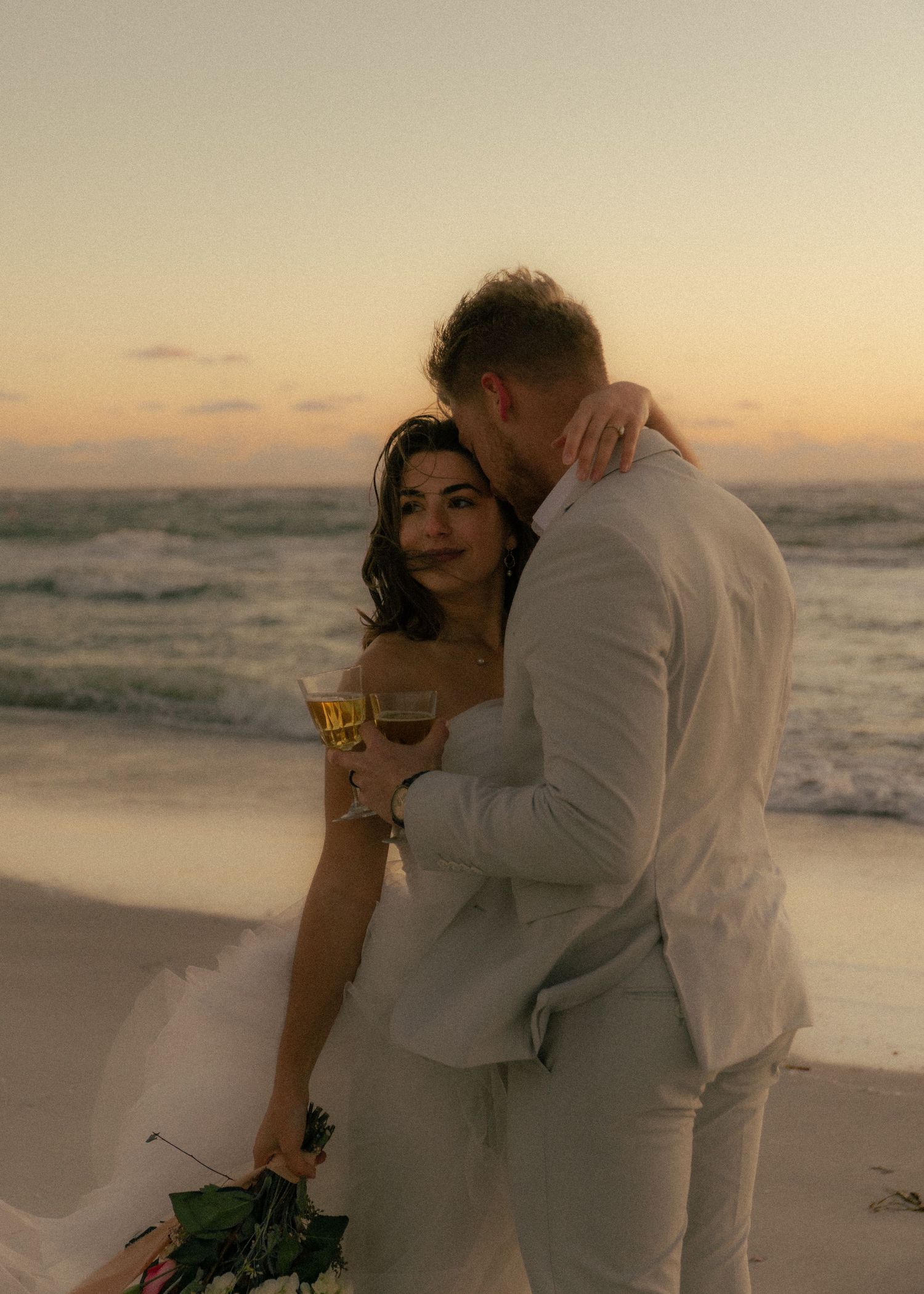 Couple in wedding attire embracing on a beach at sunset, woman holding champagne glass with ocean waves behind them.