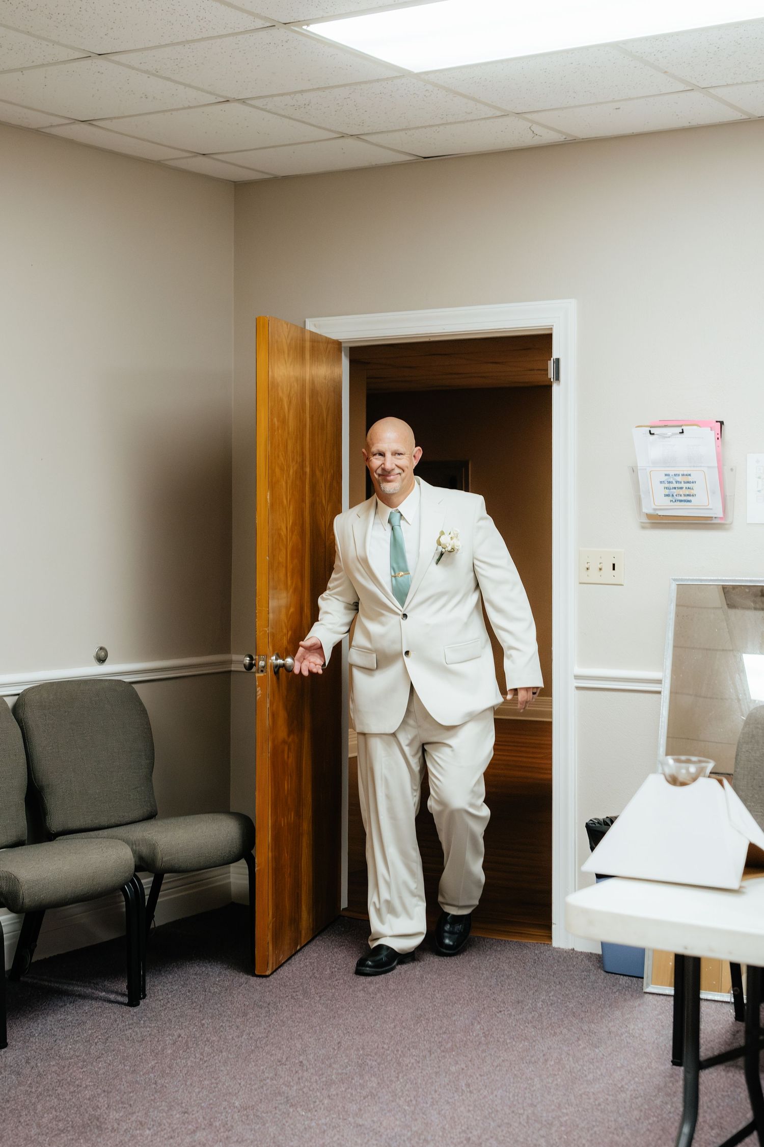 Bald groom in cream suit and mint tie smiling while opening a wooden door before his wedding ceremony.