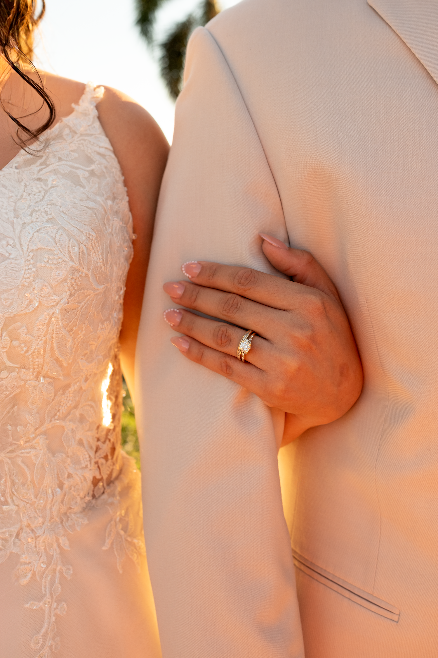 Close-up of bride's hand with diamond wedding ring resting on groom's beige suit jacket at golden hour.