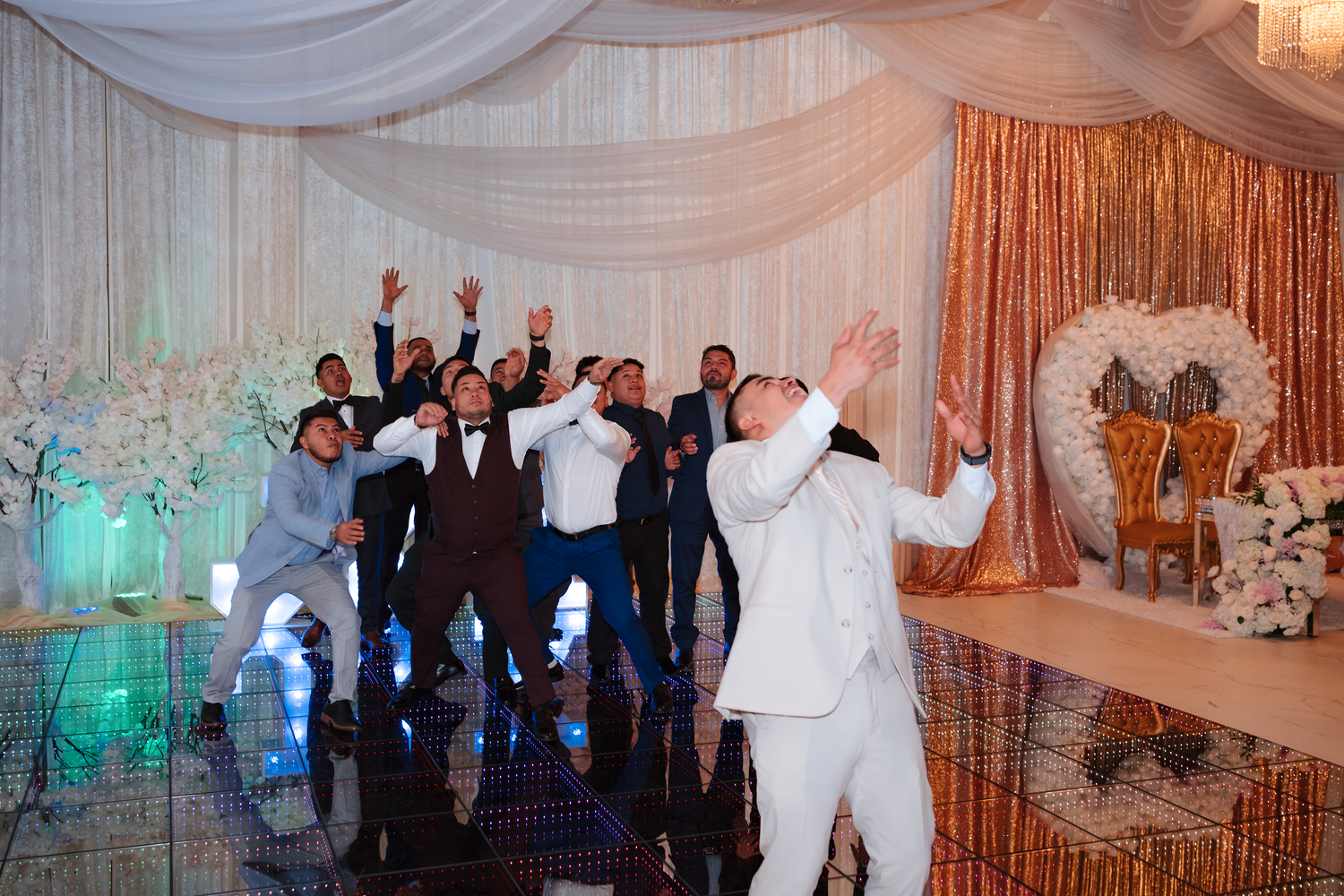 Groomsmen cheering and celebrating with raised arms on a mirrored wedding reception dance floor.