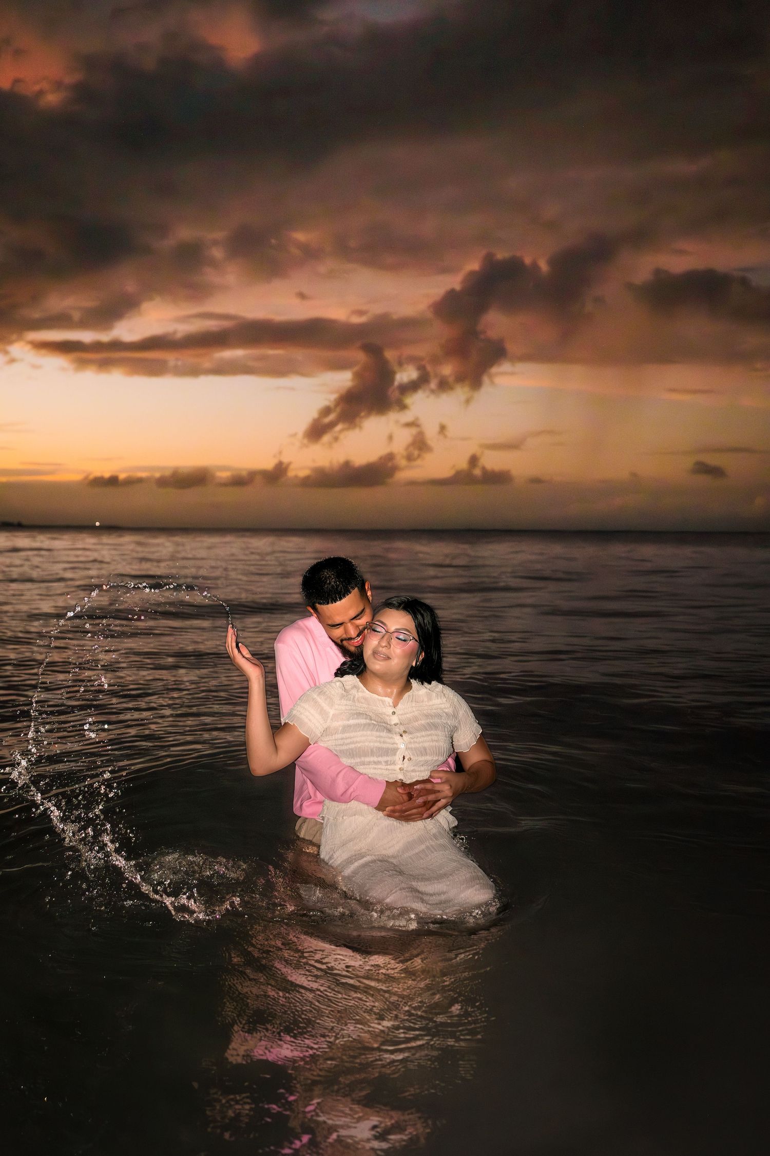 A couple embraces in dark ocean waters during sunset as dramatic storm clouds gather overhead.