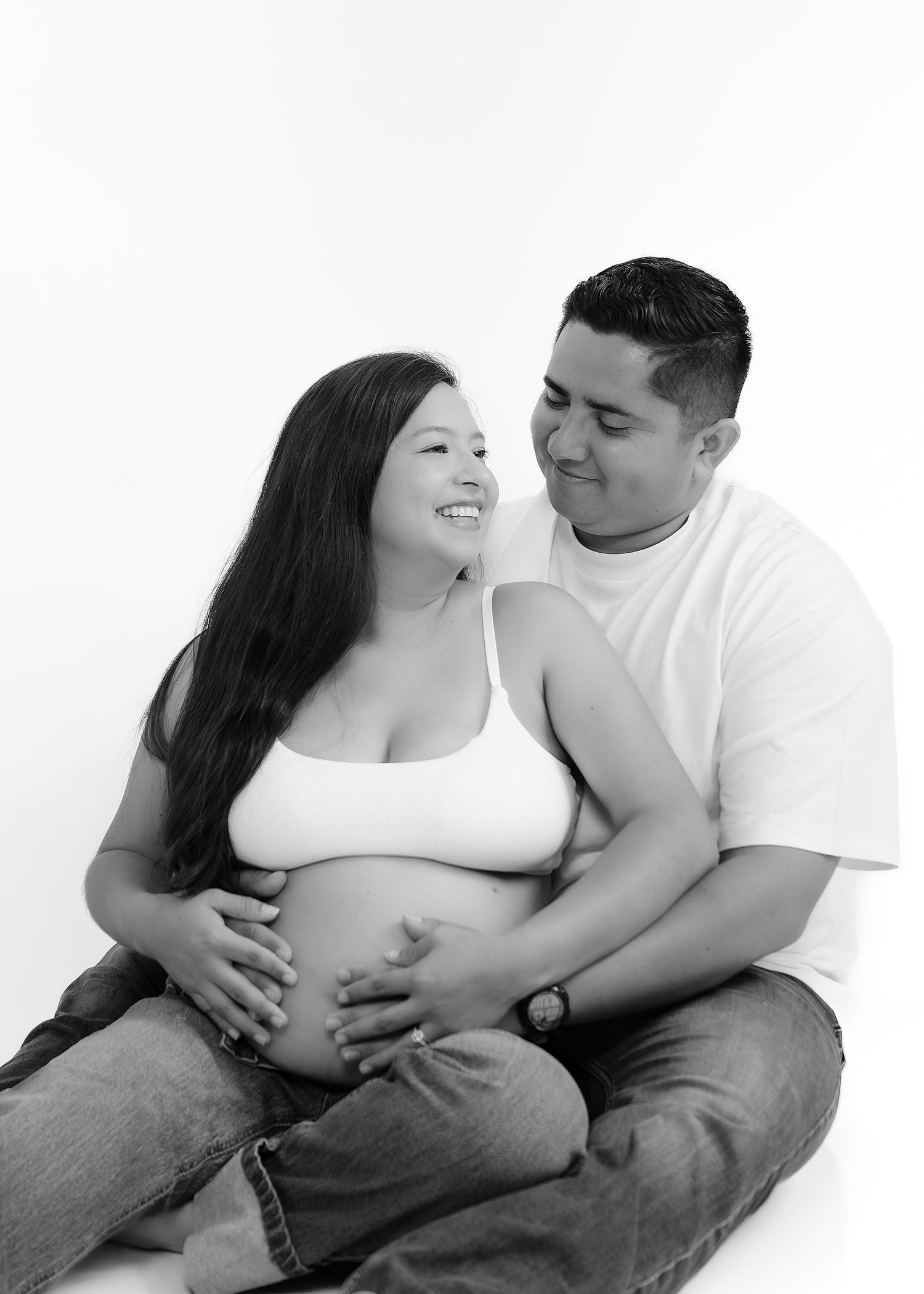 Black and white maternity photo of a smiling couple sitting together, man embracing pregnant woman's belly.