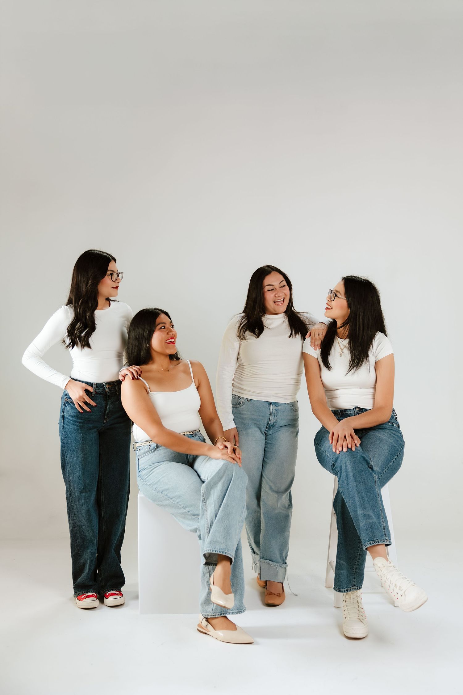 Four women in white tops and jeans pose together in a studio, smiling and interacting in a casual, friendly group photo.