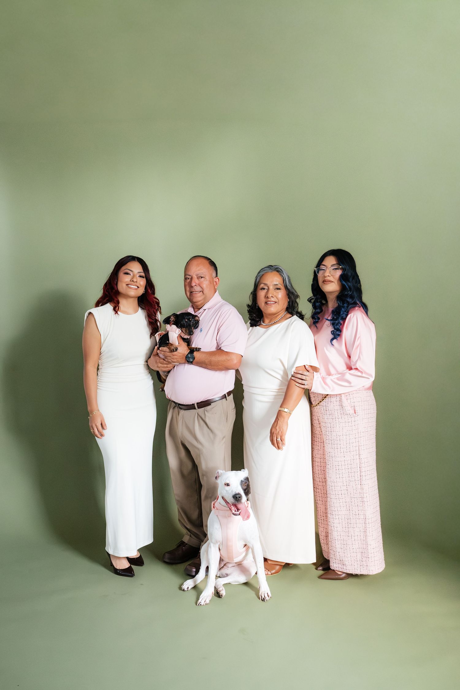 Family of four posing with two dogs against a sage green backdrop in coordinated white and pink outfits.