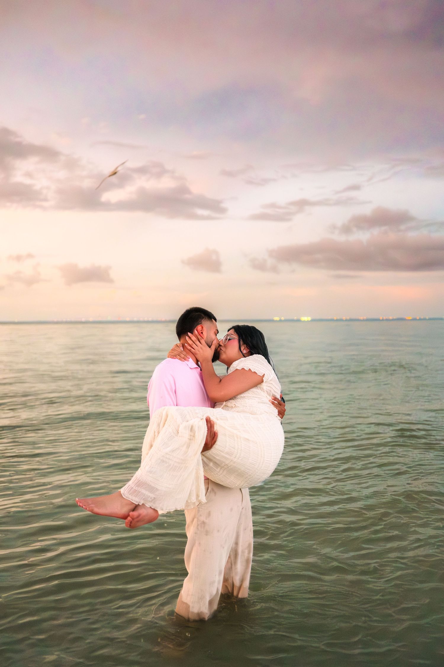 Couple kissing at sunset while standing in ocean water, man carrying woman in white dress on romantic beach.