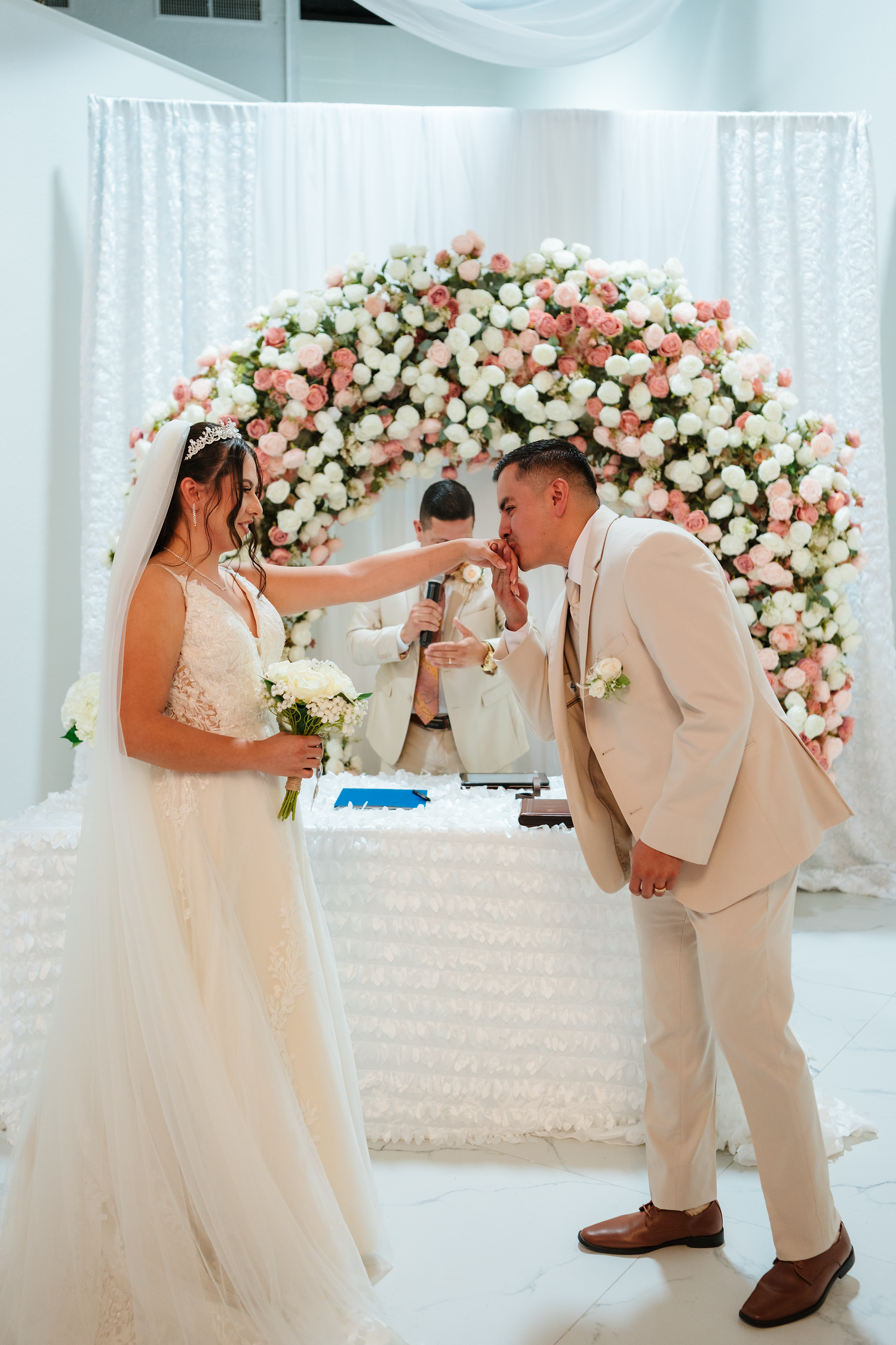 Bride and groom exchange rings at elegant wedding ceremony beneath floral arch of pink and white roses.