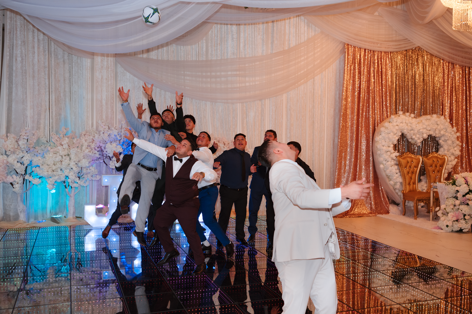 Groom and groomsmen celebrate joyfully on decorated wedding reception dance floor with gold backdrop.