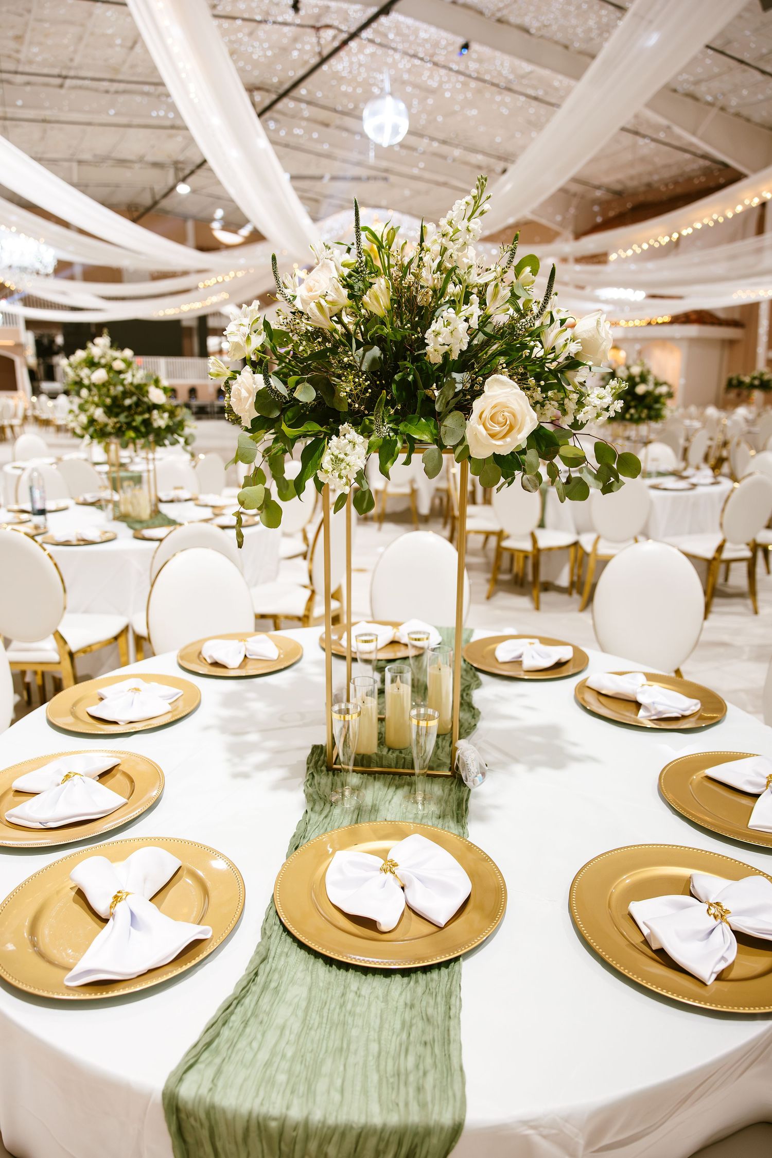 Round wedding table with gold chargers, green runner, and tall floral centerpiece.