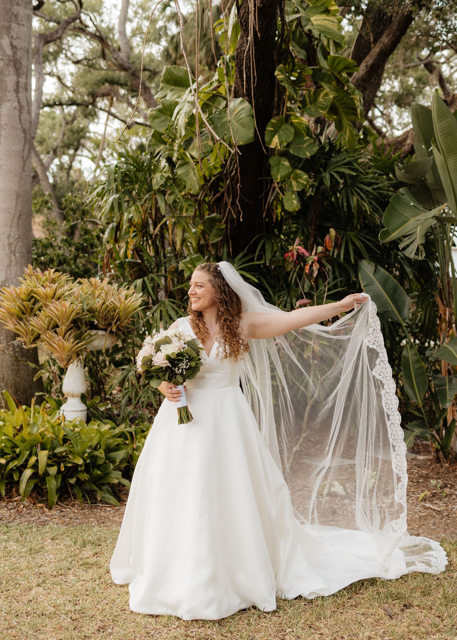 Bride in a white ball gown and lace veil poses with bouquet among lush tropical greenery.