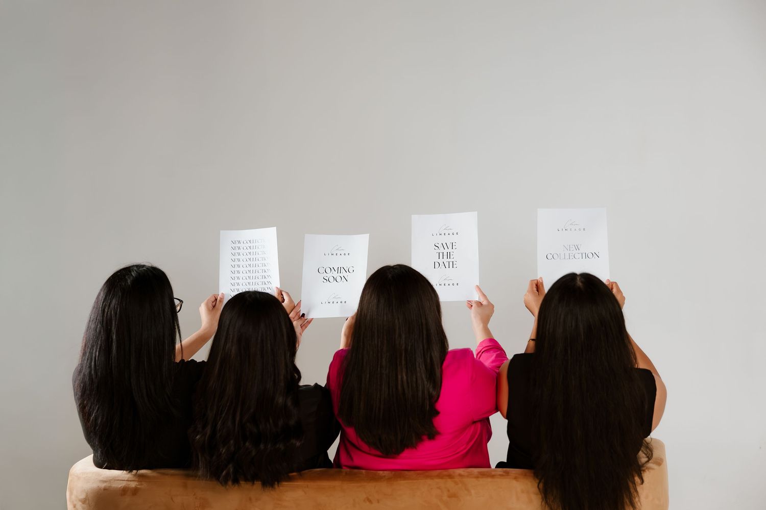 Four women with long dark hair sit together holding white papers with text, viewed from behind.