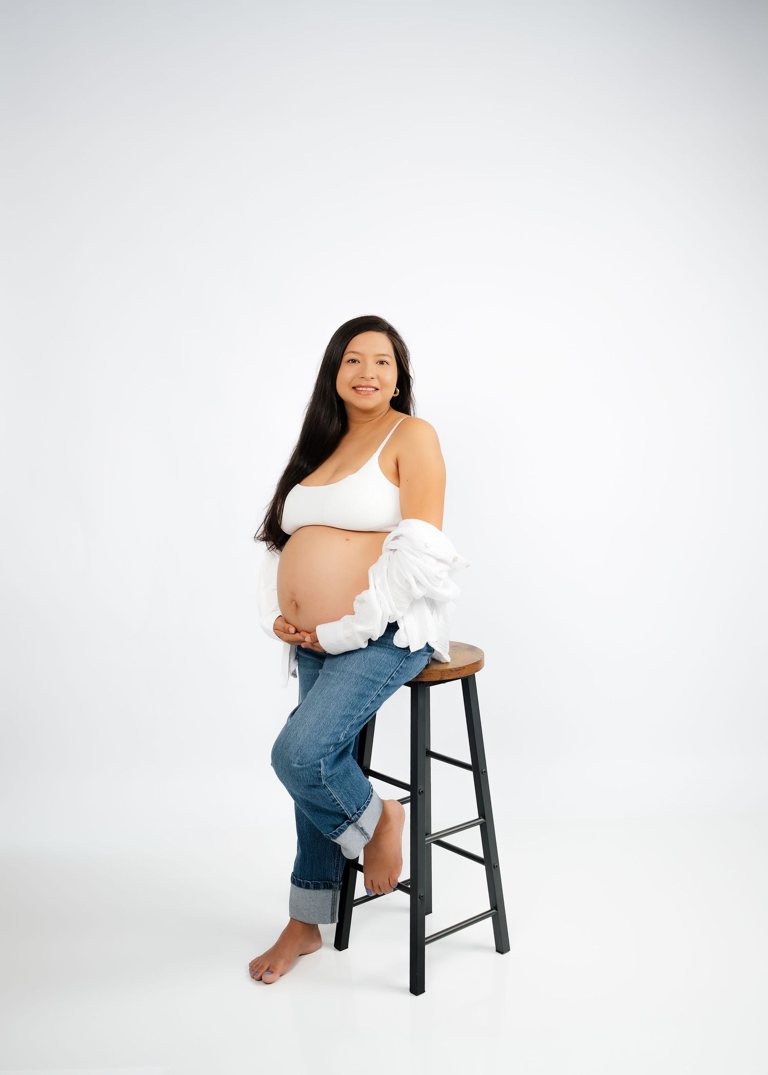 Pregnant woman in white crop top and jeans sits on wooden stool, smiling, holding baby bump in studio setting.