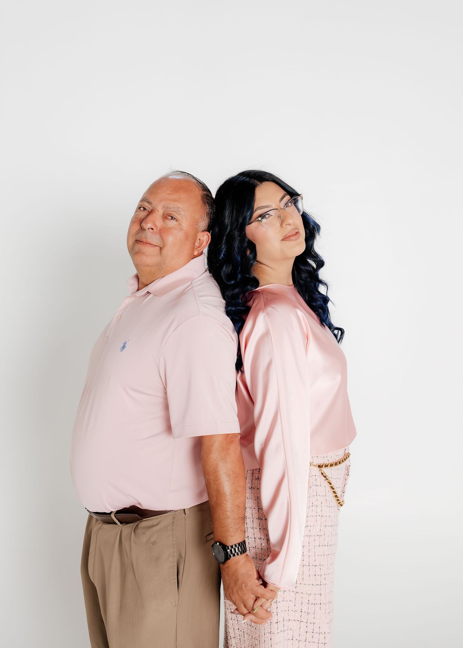 A man and woman in matching pink outfits pose back-to-back holding hands against a white background.