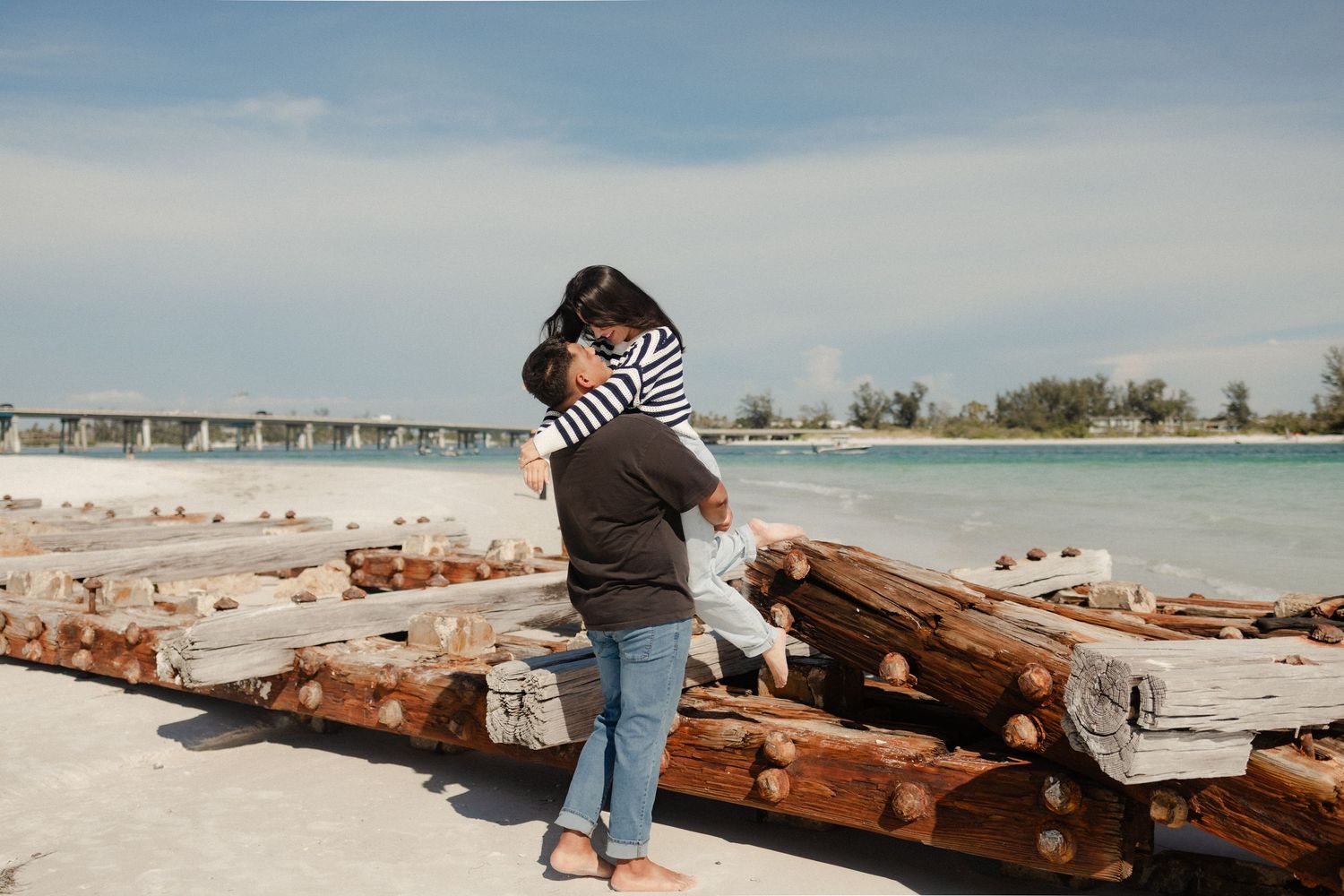 A woman hugs a child near weathered wooden logs on a sunny beach with a bridge and turquoise water in the background.