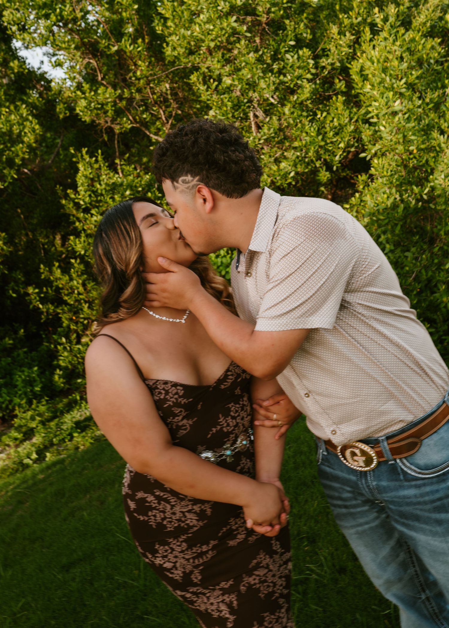 Couple sharing a romantic kiss outdoors, woman in brown floral dress and man in white polo with jeans, lush green trees behind.