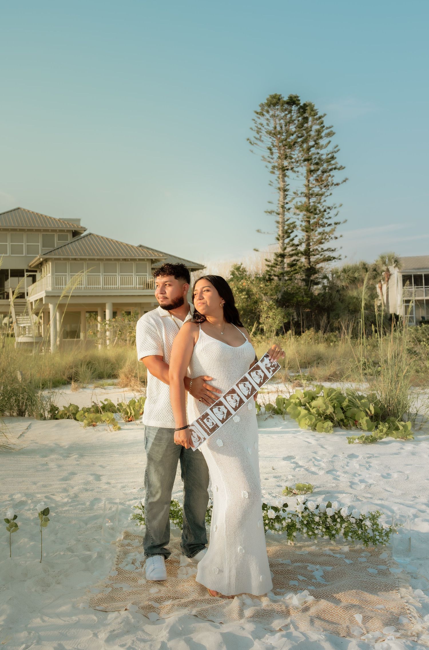 Couple poses on white sand beach at sunset, woman in white dress holding pregnancy announcement sign, man in white shirt embracing her.