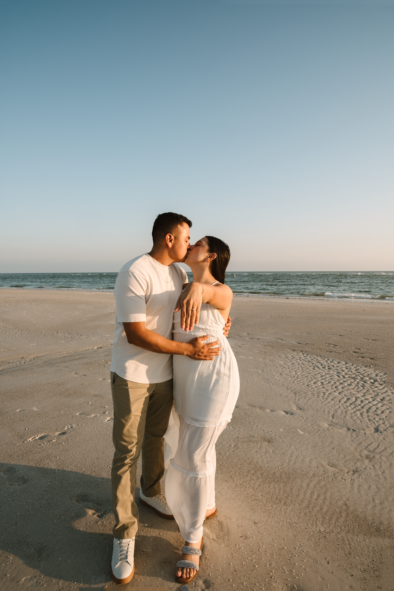 A couple shares a romantic kiss on a sandy beach at sunset, the woman in a white dress and man in a white shirt.