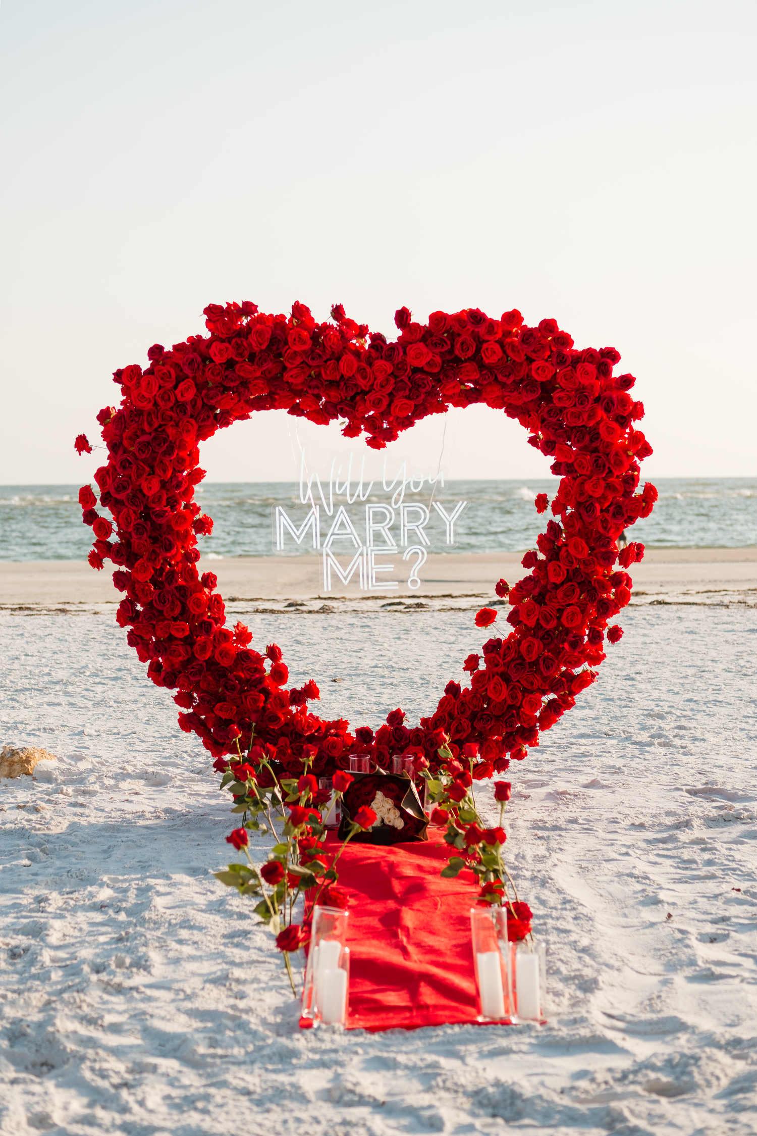 Large heart-shaped red rose arrangement on a sandy beach with a Will You Marry Me? sign in the background.