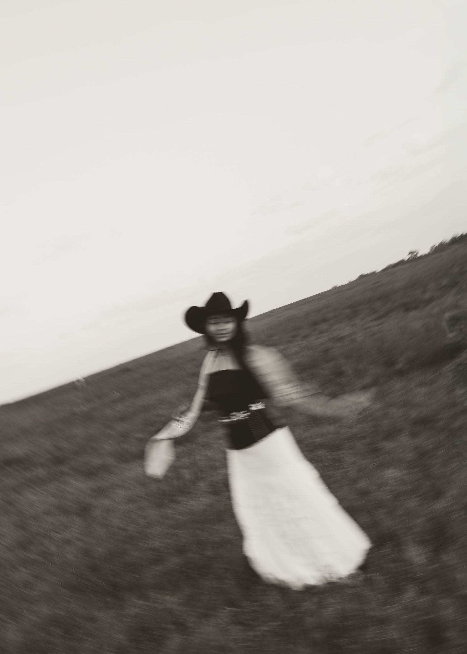 Black and white photo of a woman in a cowboy hat and white skirt dancing in an open field.