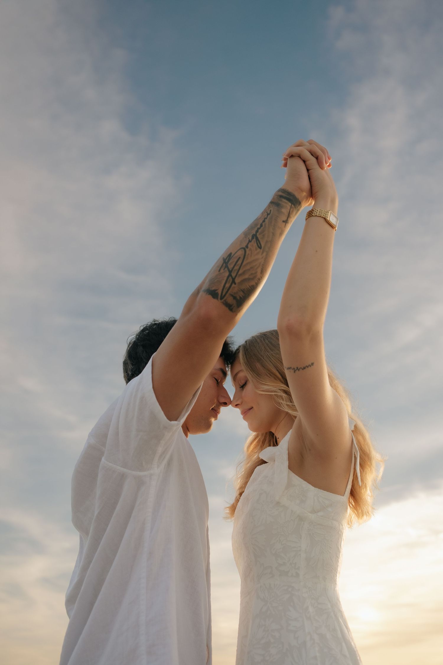 Couple in white outfits holding hands raised against a sunset sky, sharing a romantic forehead touch.