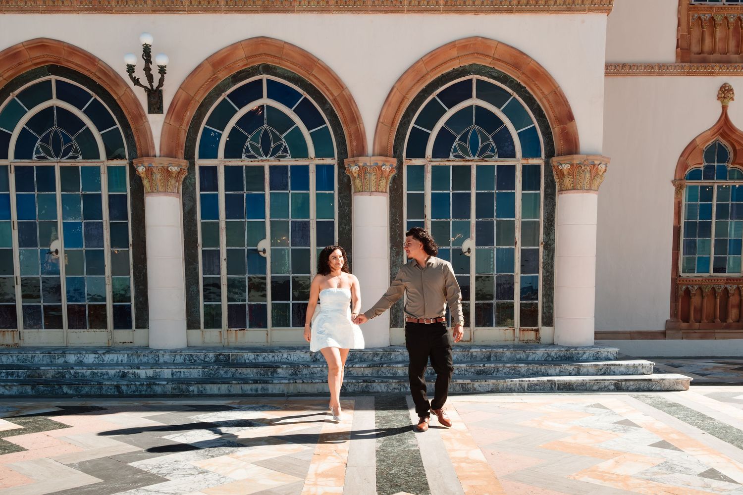 Couple holding hands walking in front of ornate building with blue stained glass arched windows on sunny day.